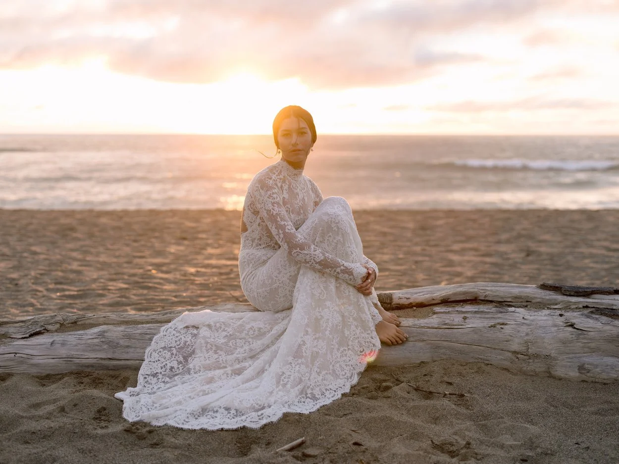 A woman in a white lace dress sitting barefoot on a driftwood log on the beach at sunset.