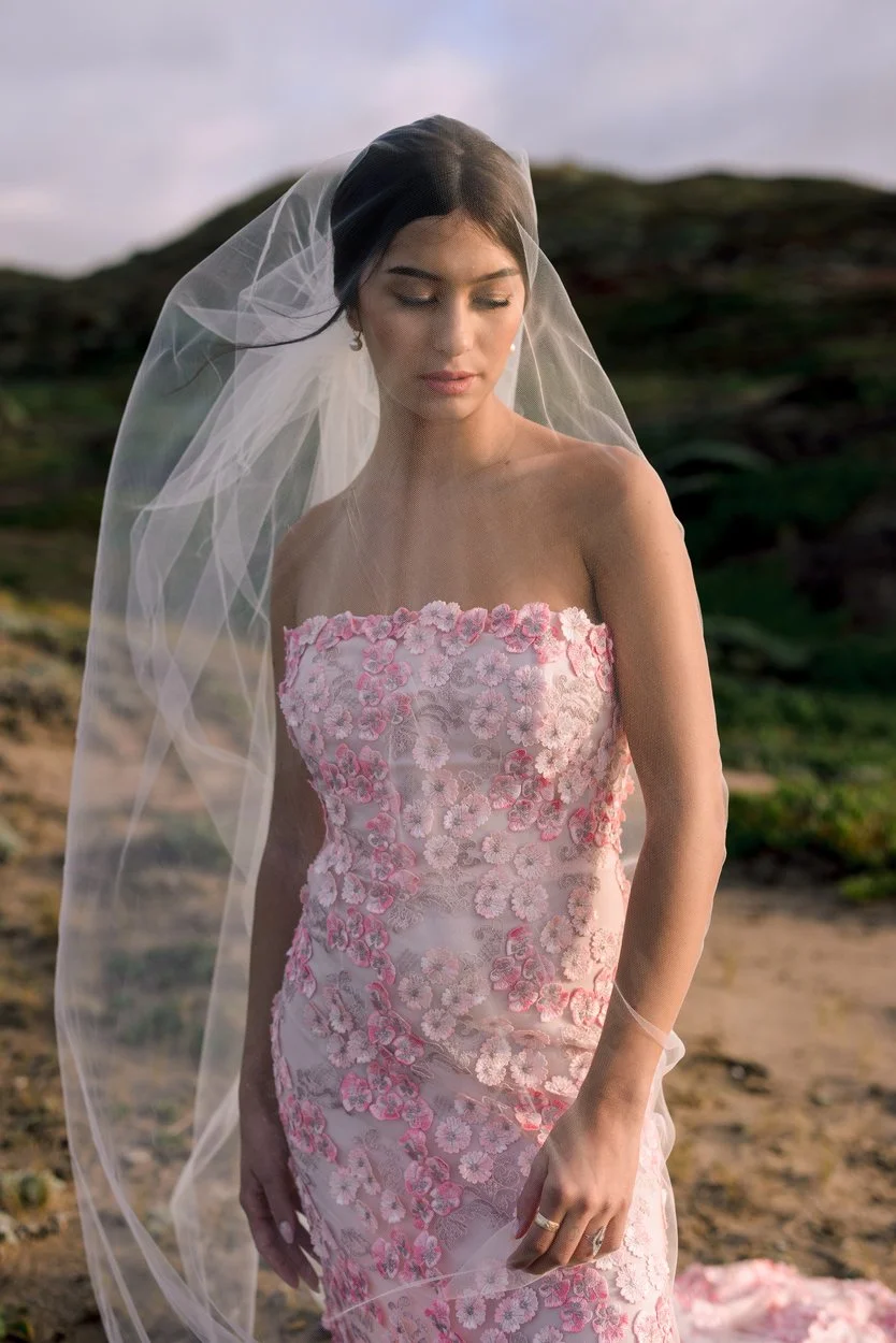 A woman in a pink floral wedding dress and veil standing outdoors with a scenic background.