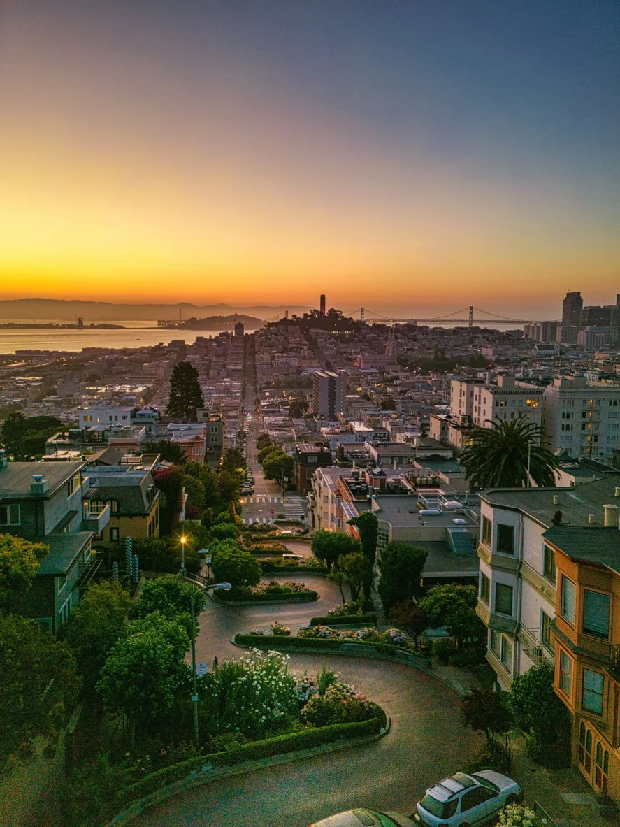 Cityscape at sunset showing a winding street, residential buildings, a hill with a tower, and a bridge over water in the distance.