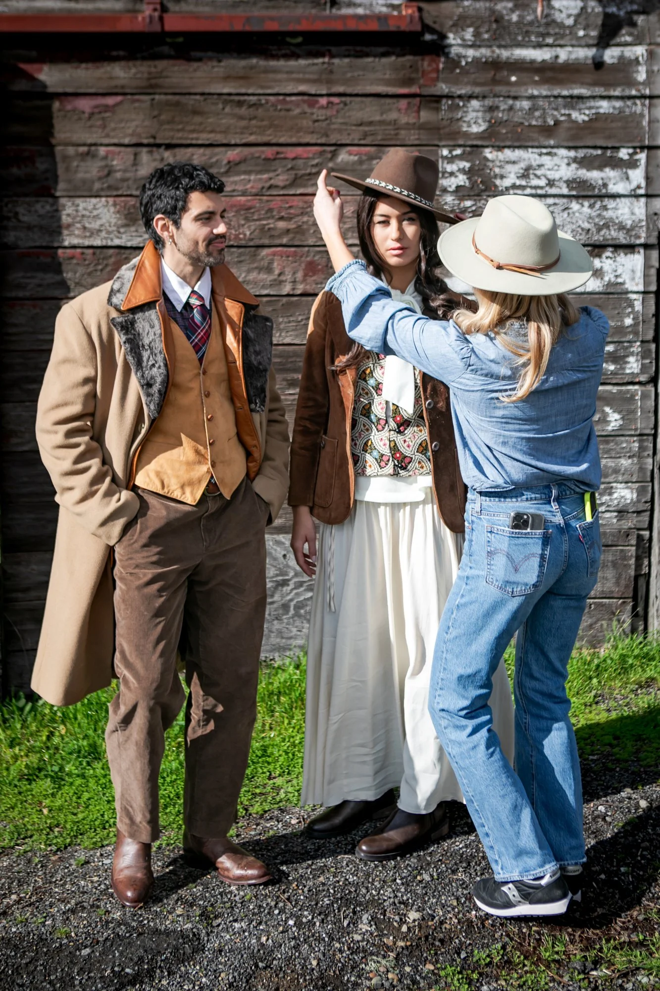Three people standing outdoors in front of a weathered wooden wall. The woman on the right is adjusting her wide-brimmed hat, while the woman in the middle is looking at her. The man on the left has his hands in his pockets and is watching them. All are dressed in vintage-style clothing with earthy tones.