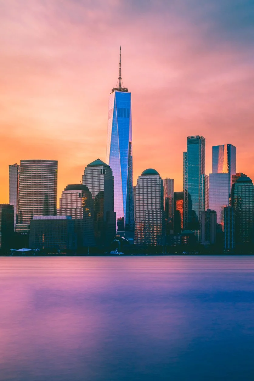 Sunset view of the New York City skyline featuring the One World Trade Center, with colorful sky and reflections on the water.
