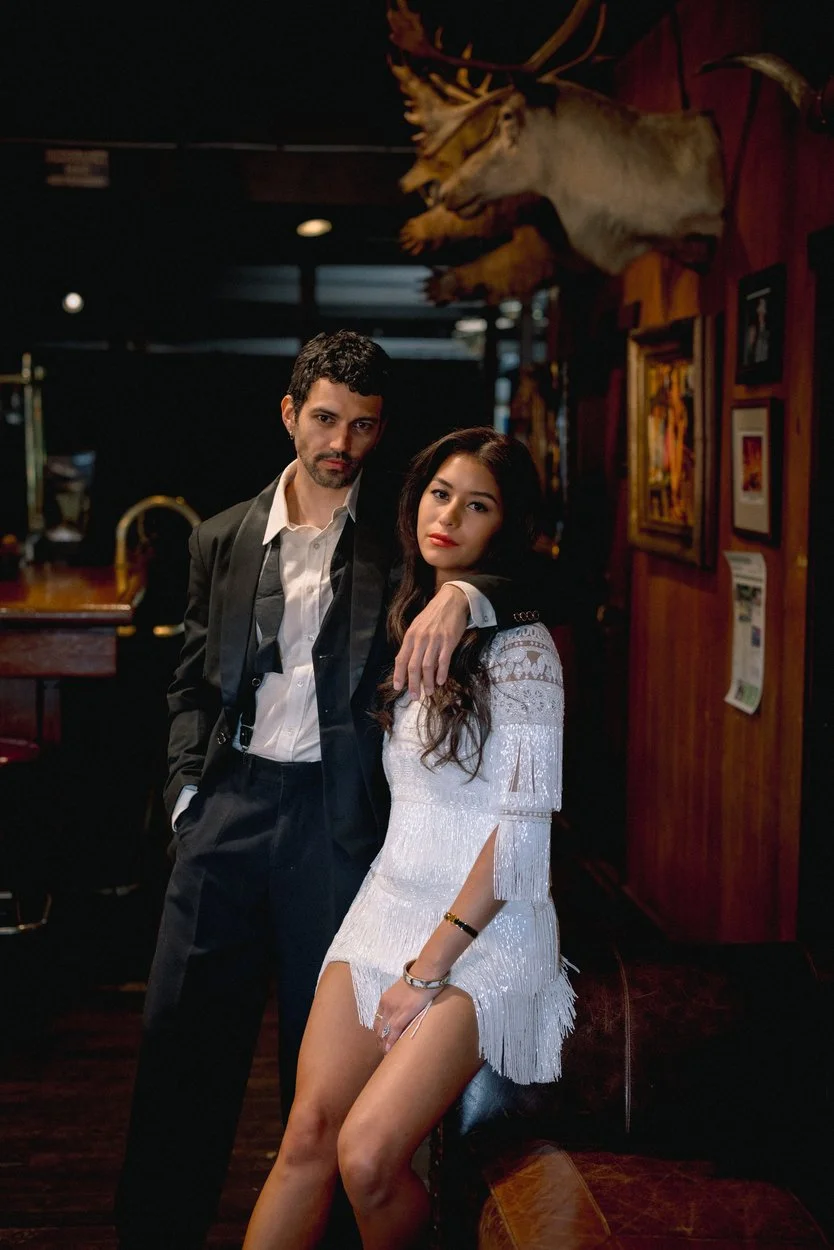 A man and woman stand together in a dimly lit bar or restaurant, with the man's arm resting on the woman's shoulder. The woman is dressed in a white, fringed dress and sitting on a stool, while the man is in a black suit with a white shirt. The backg