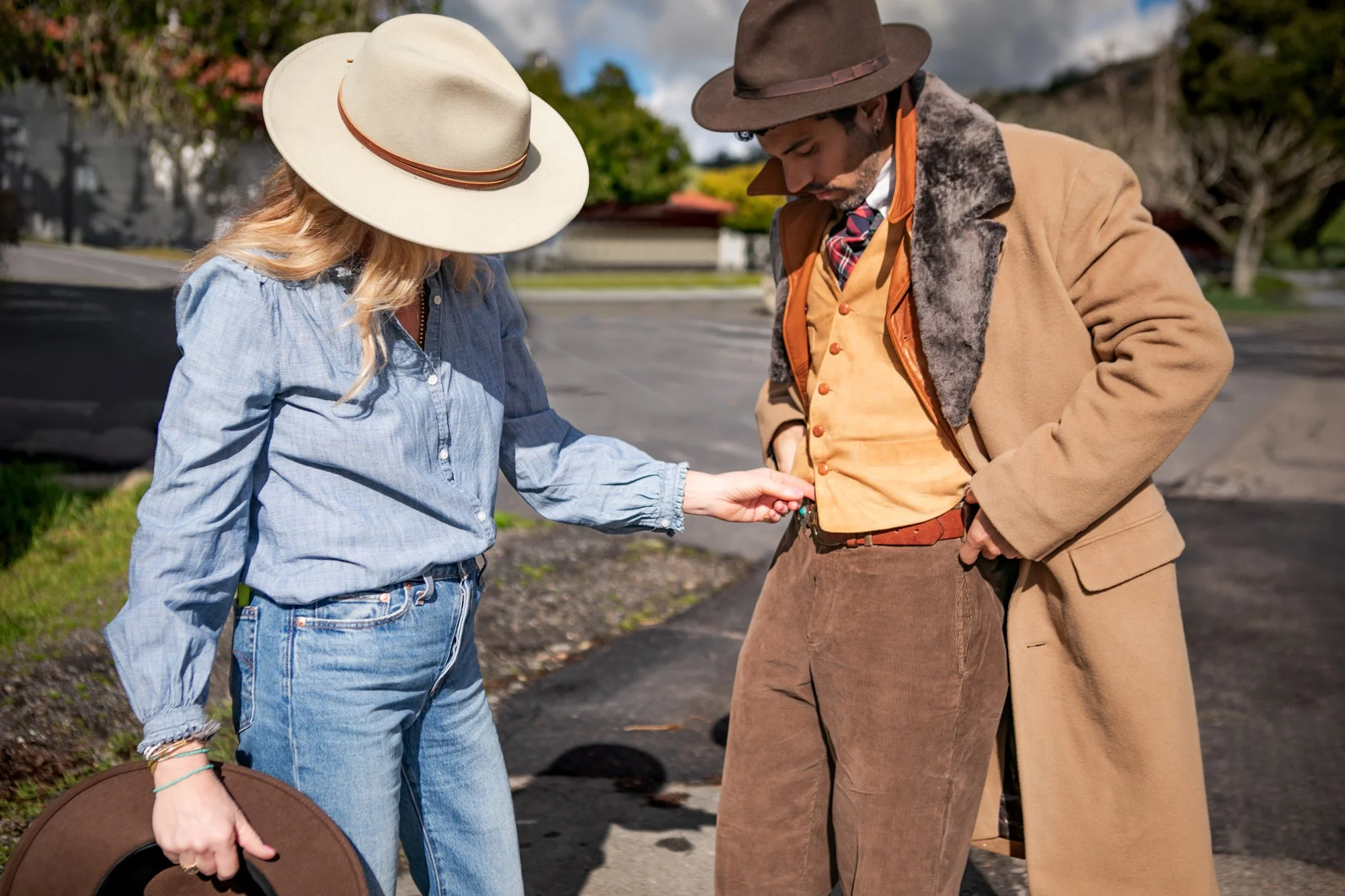 A woman and a man, both dressed in vintage-style clothing, are standing outdoors. The woman, wearing a large beige hat and a light blue shirt, is holding a brown hat in her left hand and appears to be adjusting or inspecting the man's waistband. The man, dressed in a brown hat, a tan coat with faux fur collar, and a colorful checkered scarf, is looking down at his waistband. The background shows a parking lot, trees, and a cloudy sky.