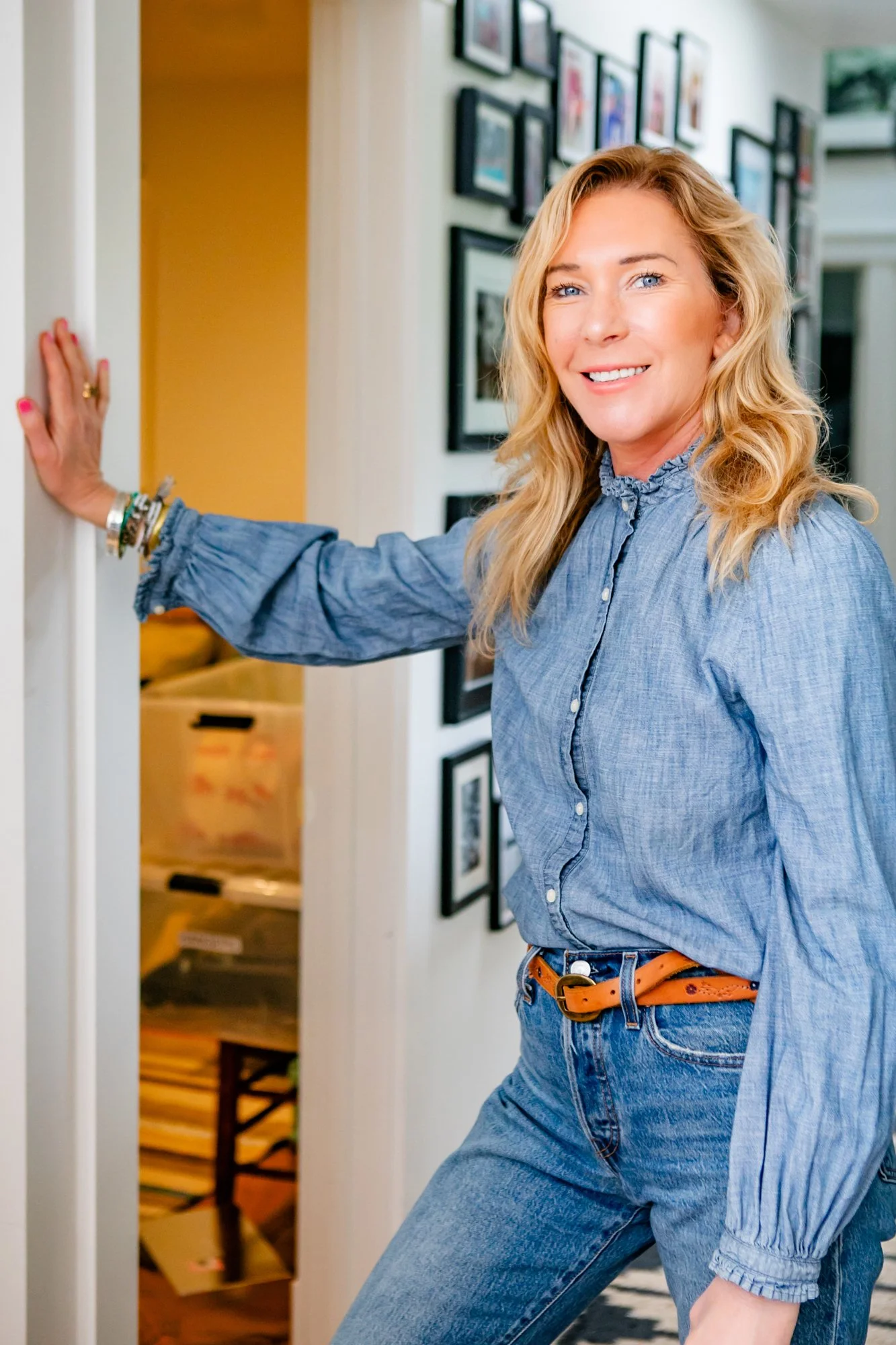 A woman with blonde hair, wearing a blue shirt and jeans, standing in a hallway with framed pictures on the wall, smiling and holding a door frame.