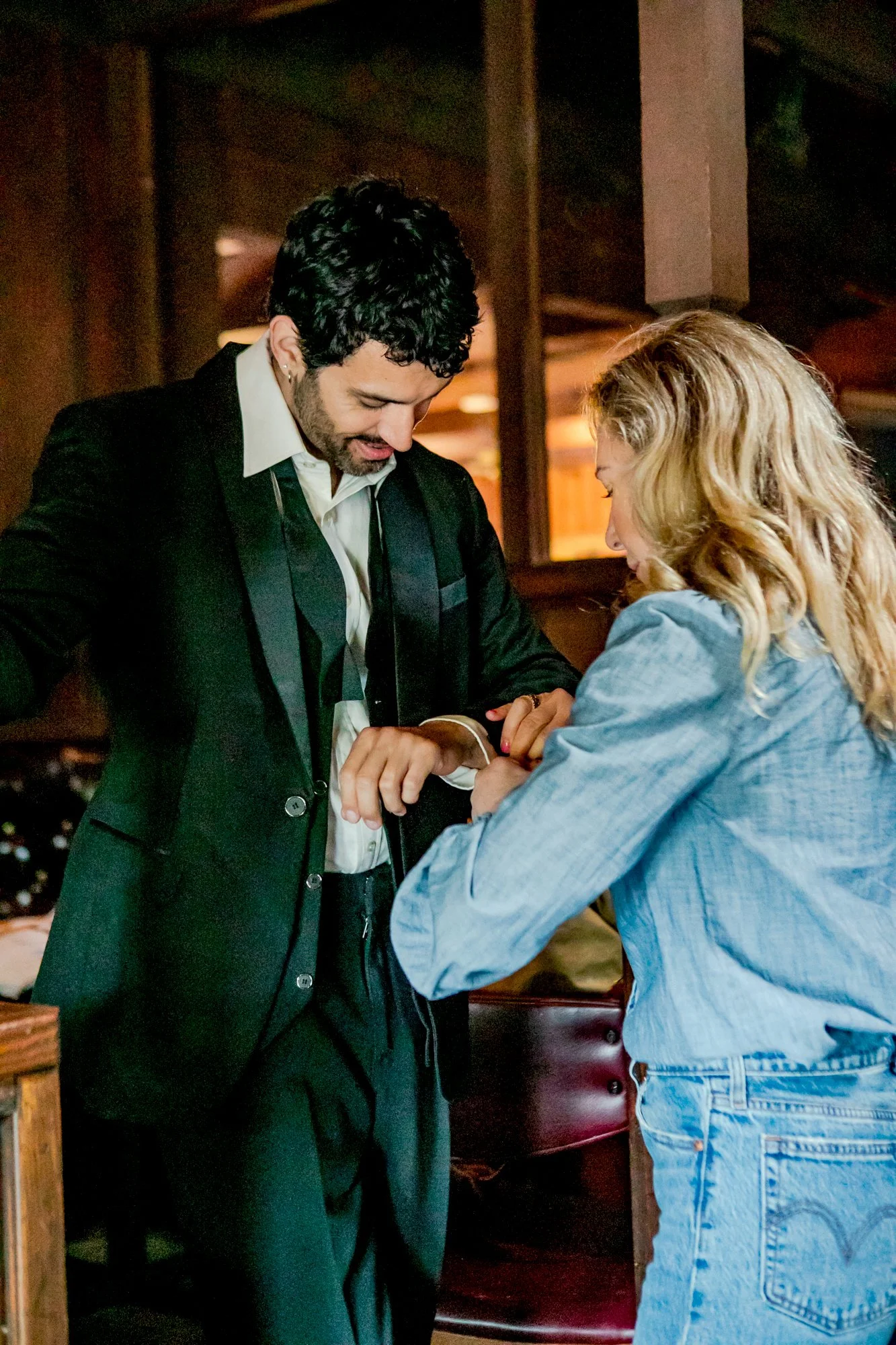 A man and a woman are standing in a cozy restaurant, engaged in a moment of affection. The man has dark curly hair, wears a black suit with a white shirt, and smiles softly at the woman. The woman has long blonde hair, wears a light blue denim jacket and jeans, and is gently holding and looking at the man's hands, smiling.