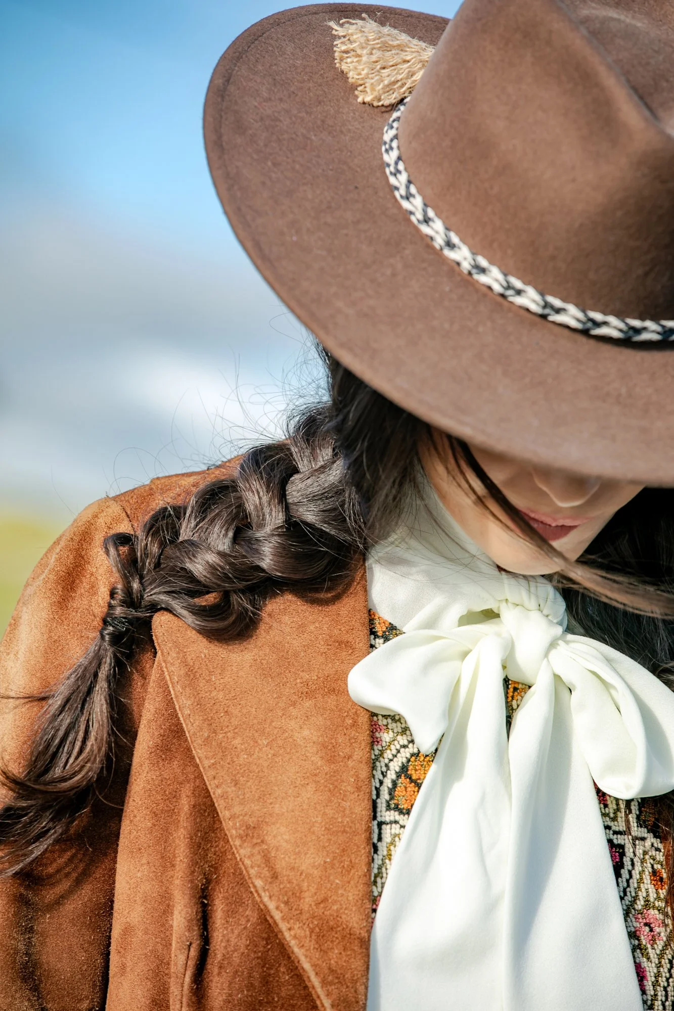 A woman wearing a brown cowboy hat with a braided hair falling over her shoulder, a brown suede jacket, a patterned shirt, and a large cream-colored bow tie.