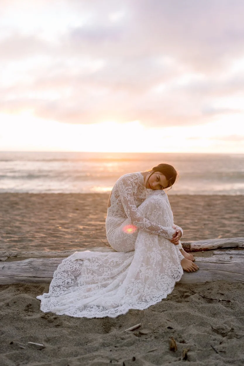 A woman in a white lace dress sitting on a beach at sunset, hugging her knees and looking at the camera.