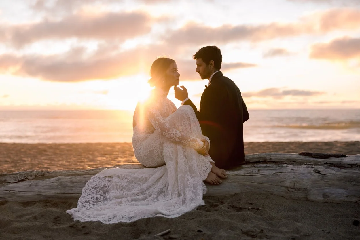 A couple, dressed in wedding attire, sitting on a log on the beach during sunset, looking at each other.