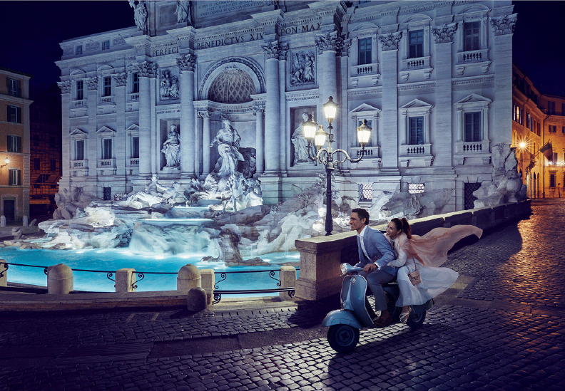 A couple riding a blue scooter at night near the Trevi Fountain in Rome, Italy, with illuminated historical buildings in the background.