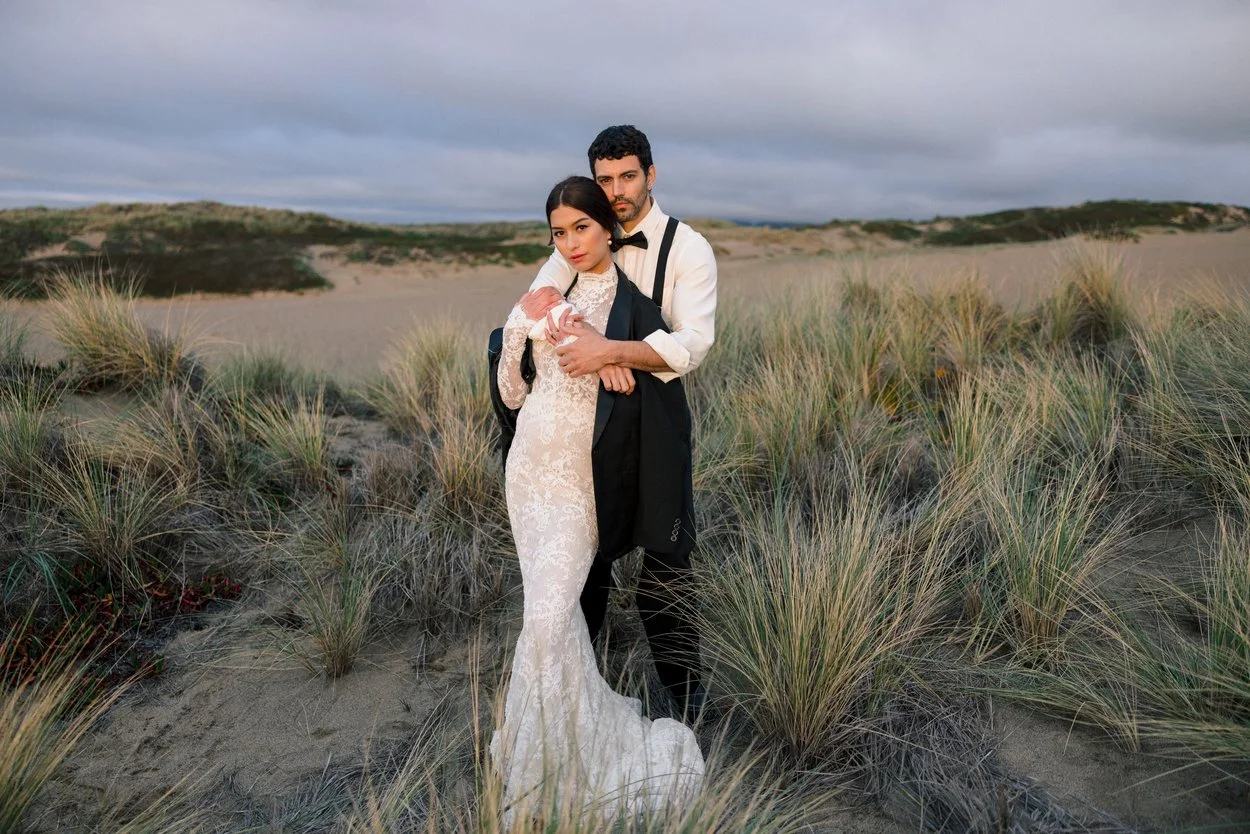 A couple dressed in formal wedding attire standing on a sandy beach with tall grass, under an overcast sky.
