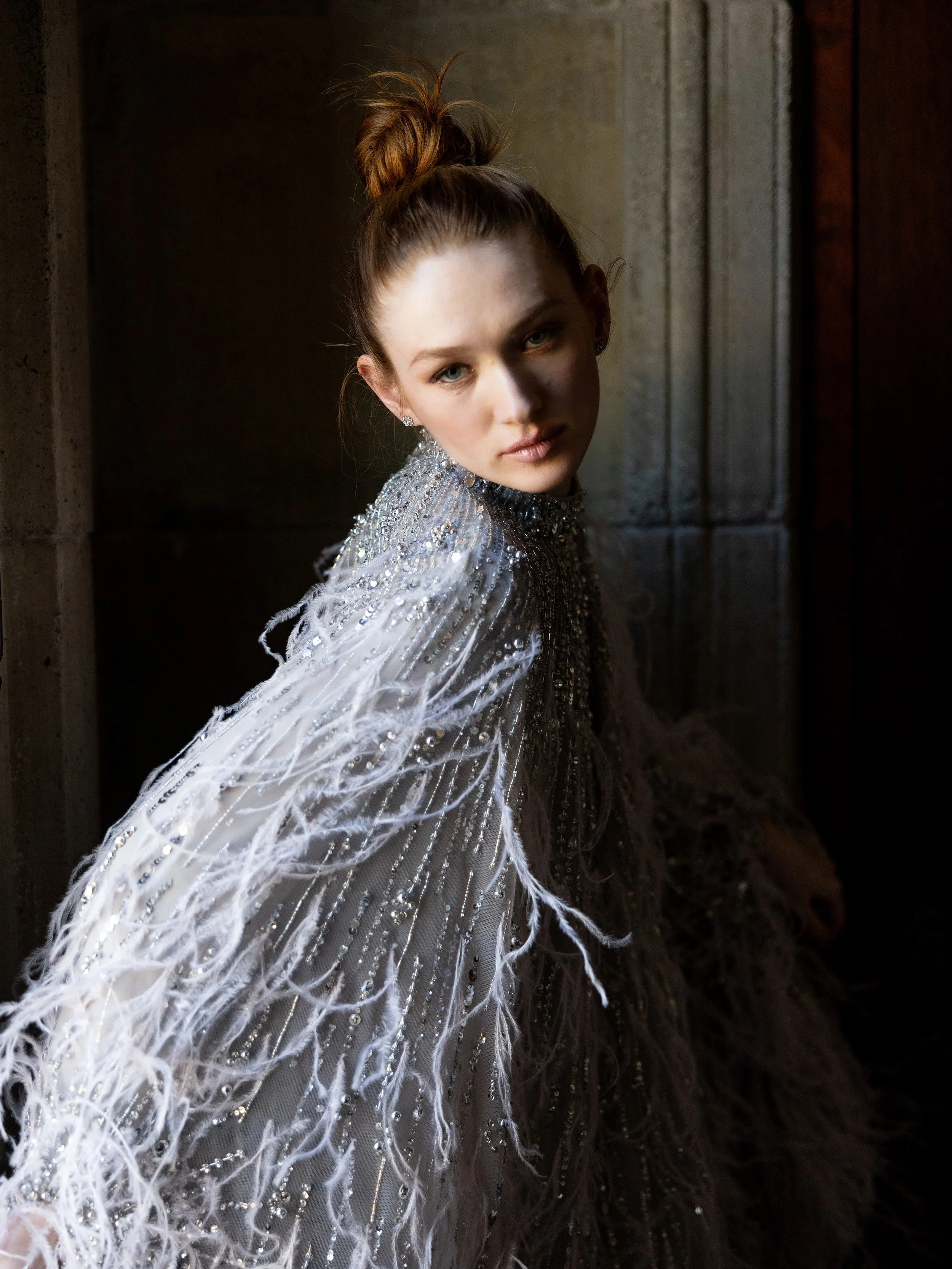 A woman with red hair styled in a high bun wears a silver, embellished, feathered gown and looks at the camera in a dimly lit room with dark walls.