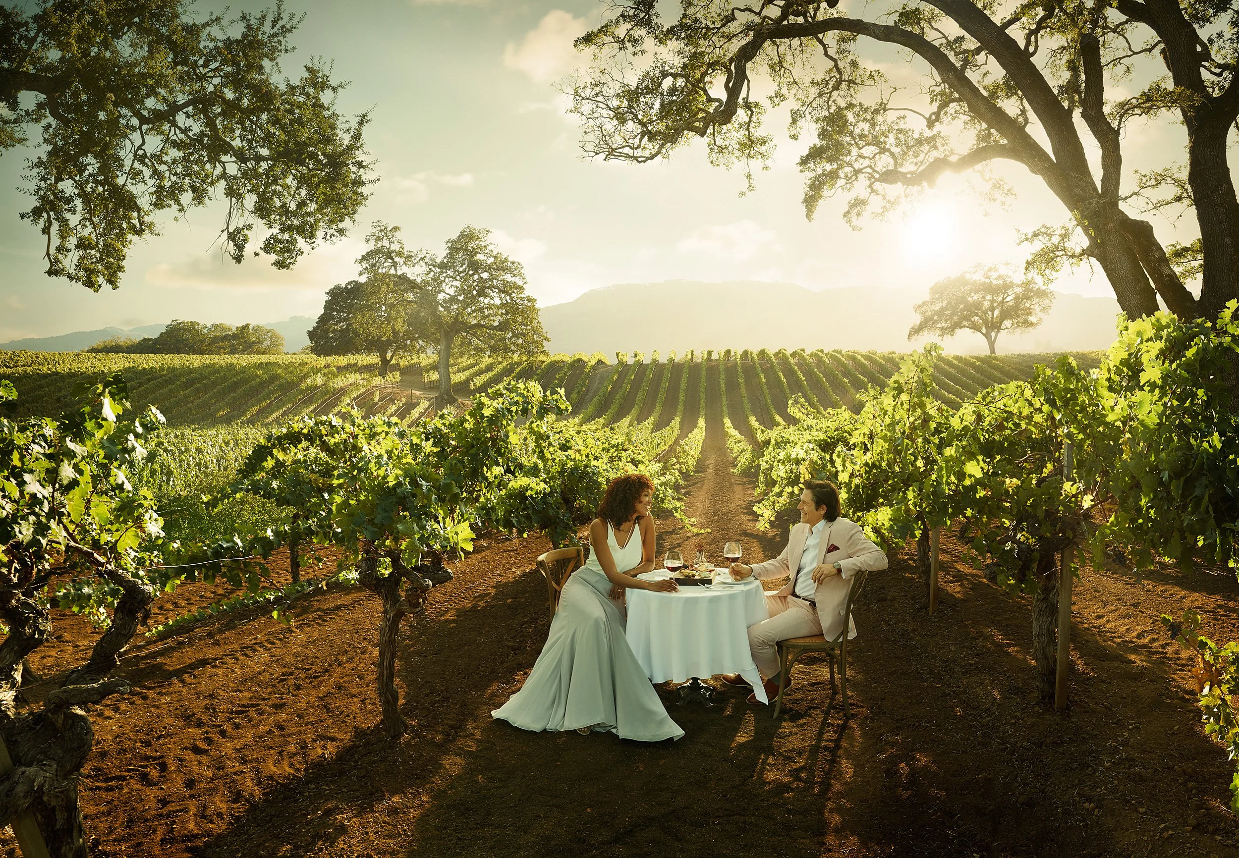 A man and woman dressed in formal clothes sitting at a small table with a white tablecloth in a vineyard during sunset, enjoying wine and conversation. The vineyard has rows of green grapevines and large trees in the background with the sun shining t