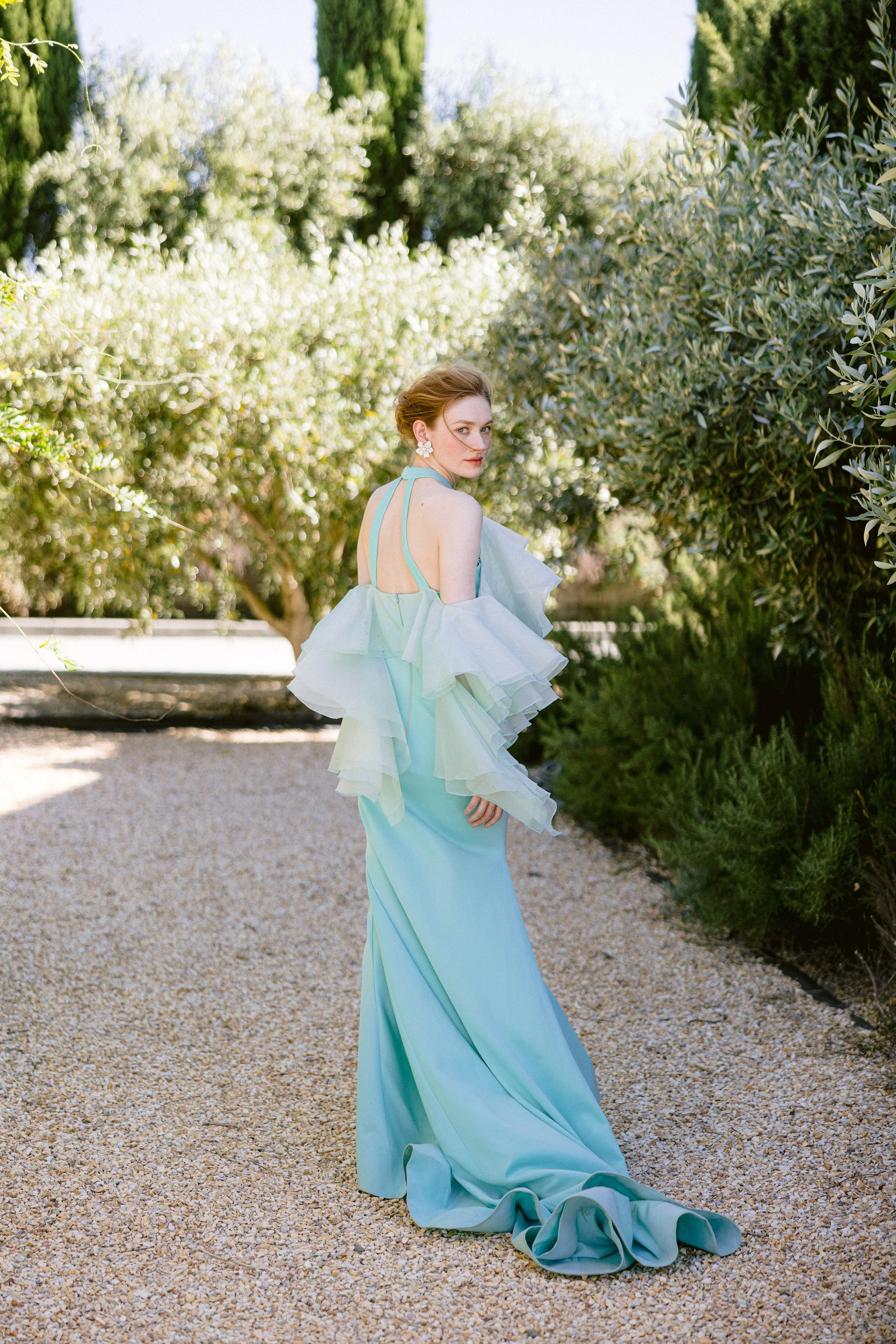 A woman in a flowing, light blue gown with ruffled sleeves, standing on a gravel path in a lush garden with trees and greenery.