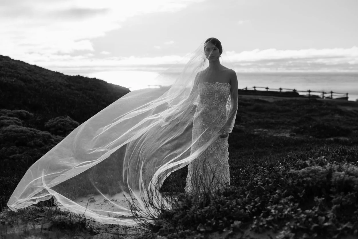 A woman in a wedding dress and veil standing outdoors on a grassy landscape during sunset or sunrise, with hills in the background.