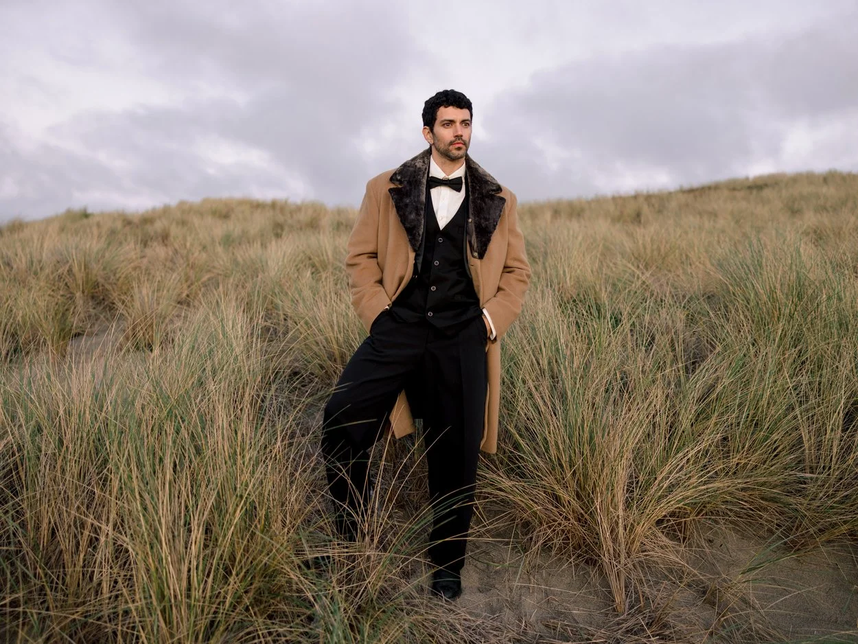 A man dressed in vintage formal attire with a long brown coat, black vest, and tie stands in a grassy field under cloudy skies.