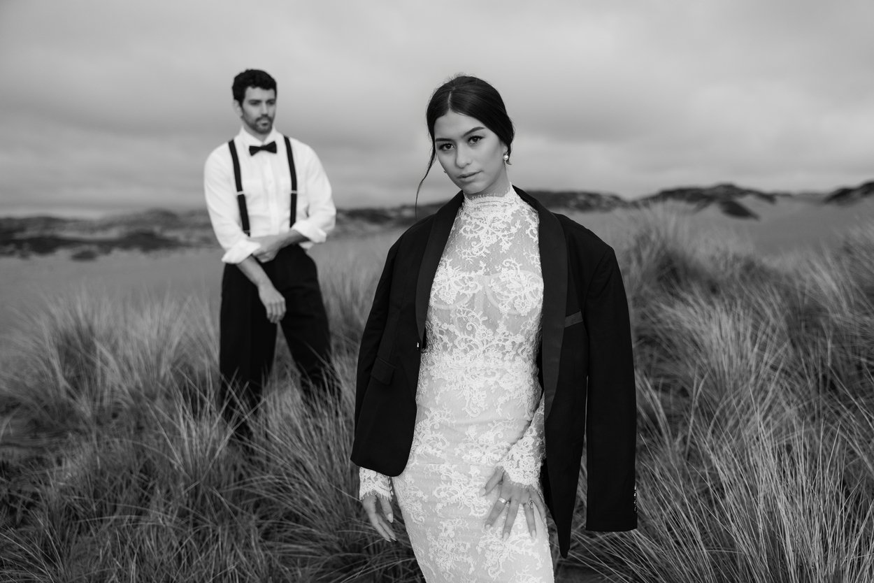 A black and white photo of a woman in a lace dress and blazer, standing in tall grass with a man in tuxedo and suspenders in the background on a scenic outdoor landscape.