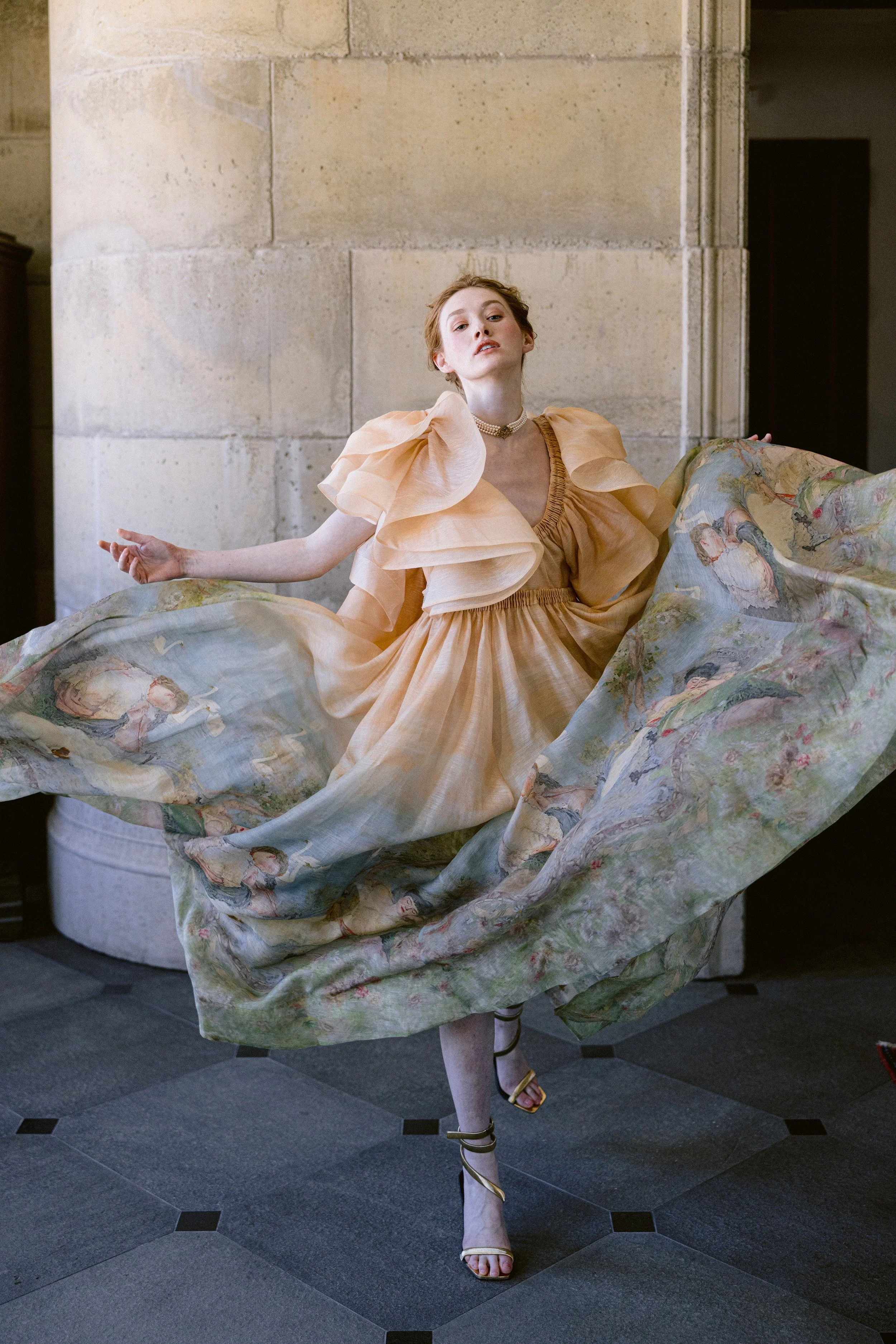 A woman in a peach-colored dress with puffed sleeves and ruffled details is standing with one leg raised, holding fabric that has a floral and figure print, in front of a stone wall.
