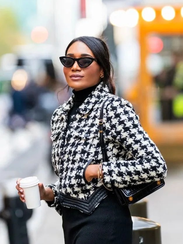 Stylish woman wearing black sunglasses, houndstooth jacket, and black pants, holding a to-go coffee cup, standing outdoors in an urban setting.