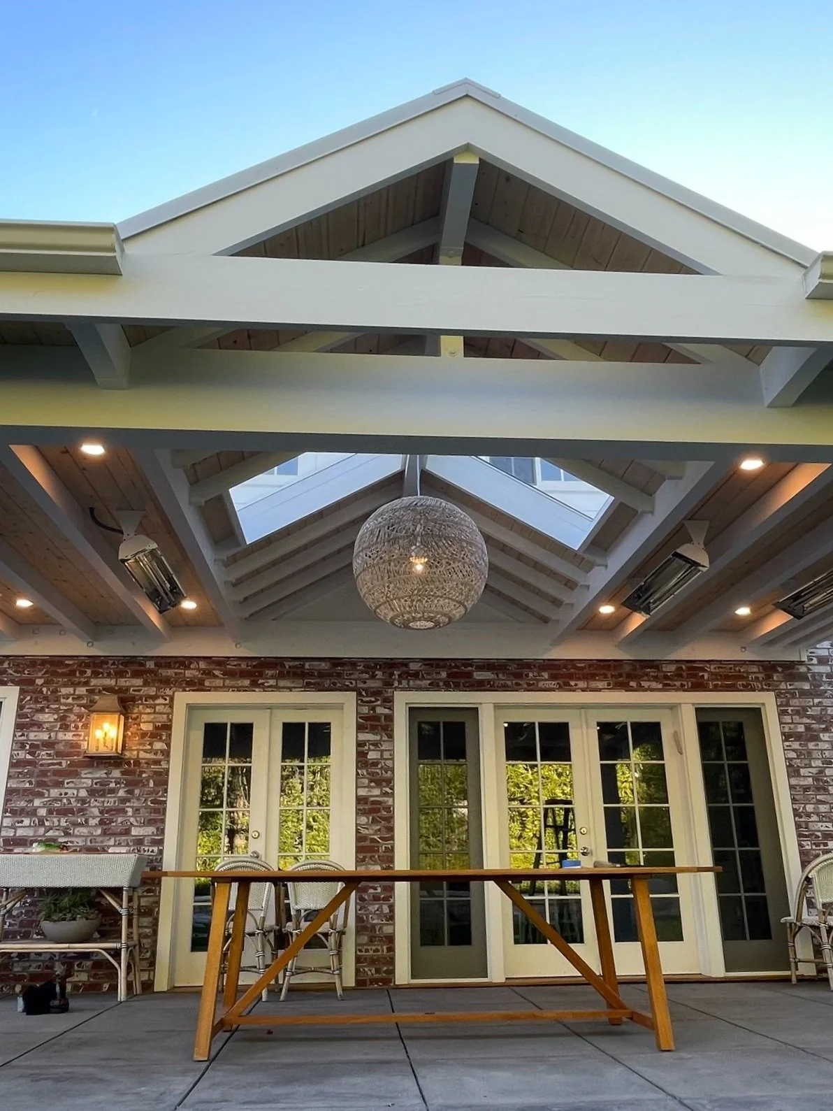 Rear view of a house with a brick exterior, double French doors, outdoor furniture, and a wooden table on a patio, with a decorative chandelier hanging from a glass-paneled ceiling, and a roof with skylights.