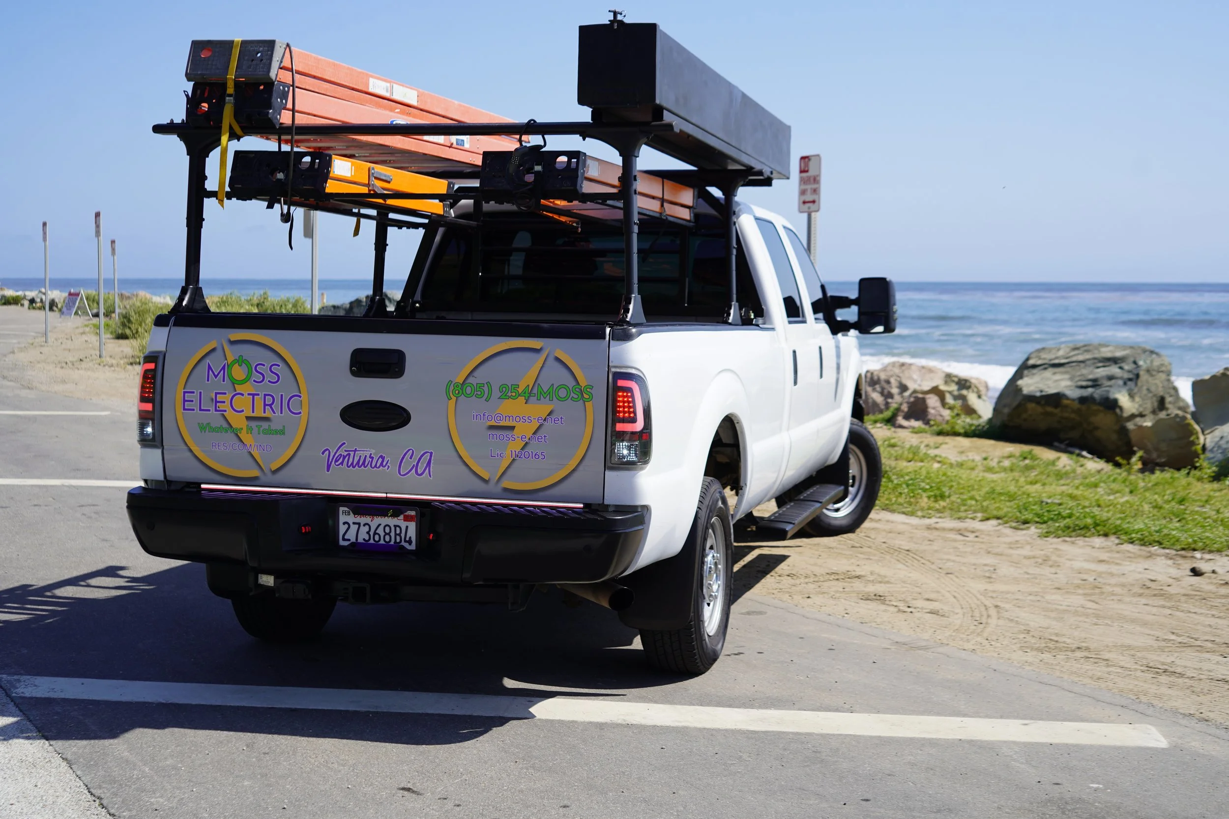 White pickup truck parked near the beach with advertising and contact information for Moss Electric on the tailgate, carrying ladders and equipment on the roof rack.