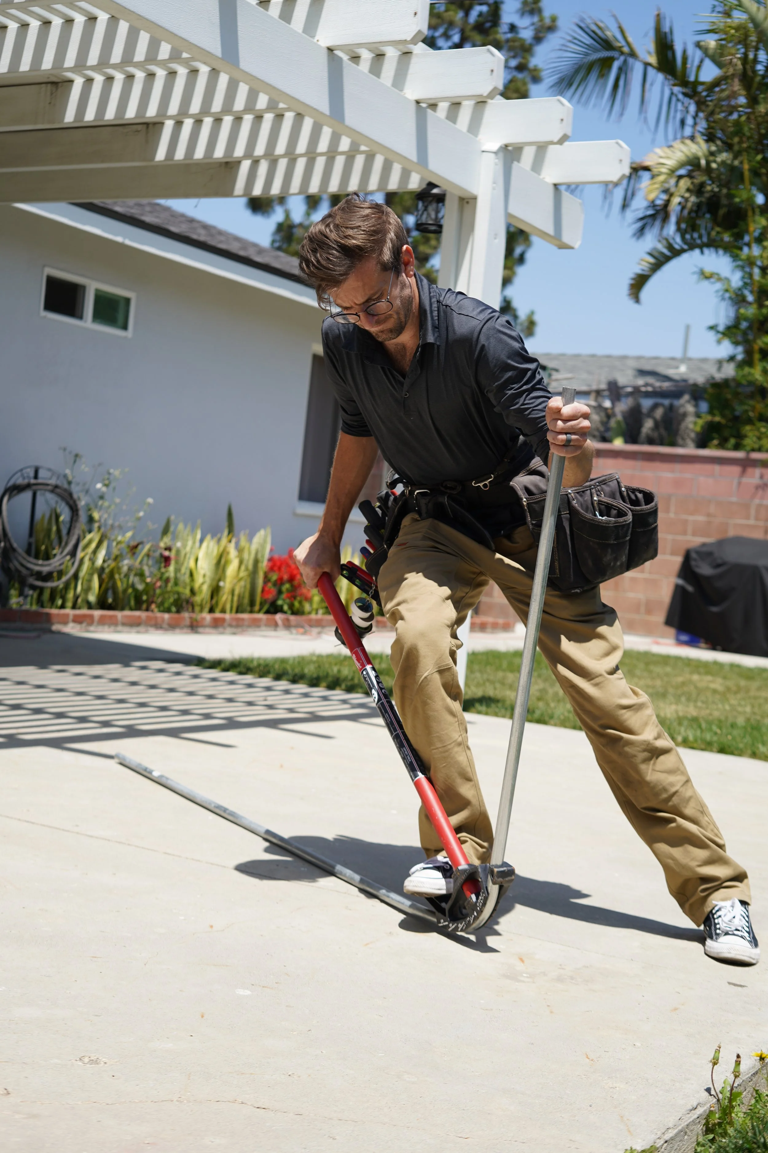 A man wearing glasses, a black shirt, and beige pants installing electrical equipment outdoors on a sunny day.