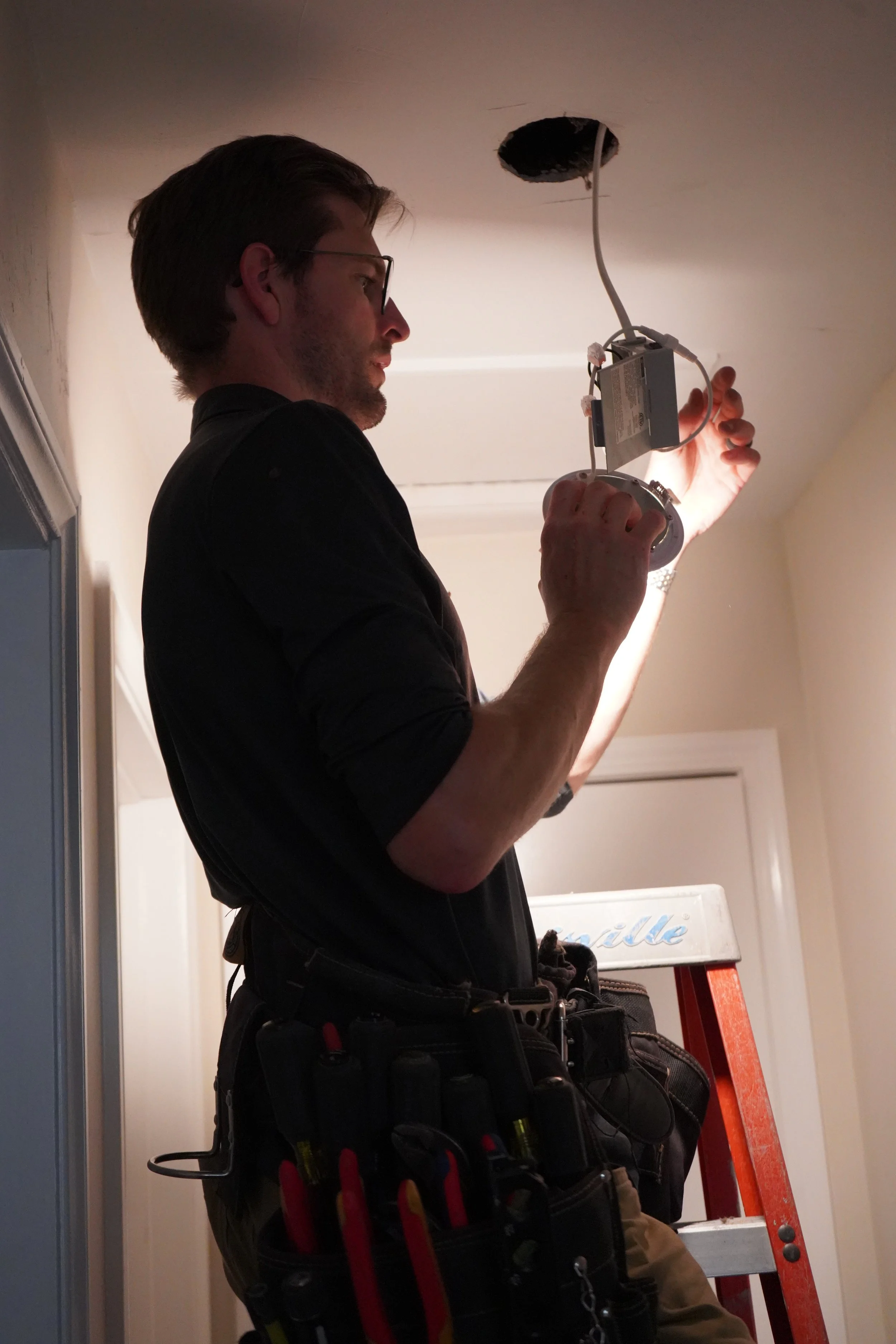 A man working on installing or repairing ceiling electrical wiring, standing on a ladder with tools around his waist, focusing on wiring connections near a ceiling hole.