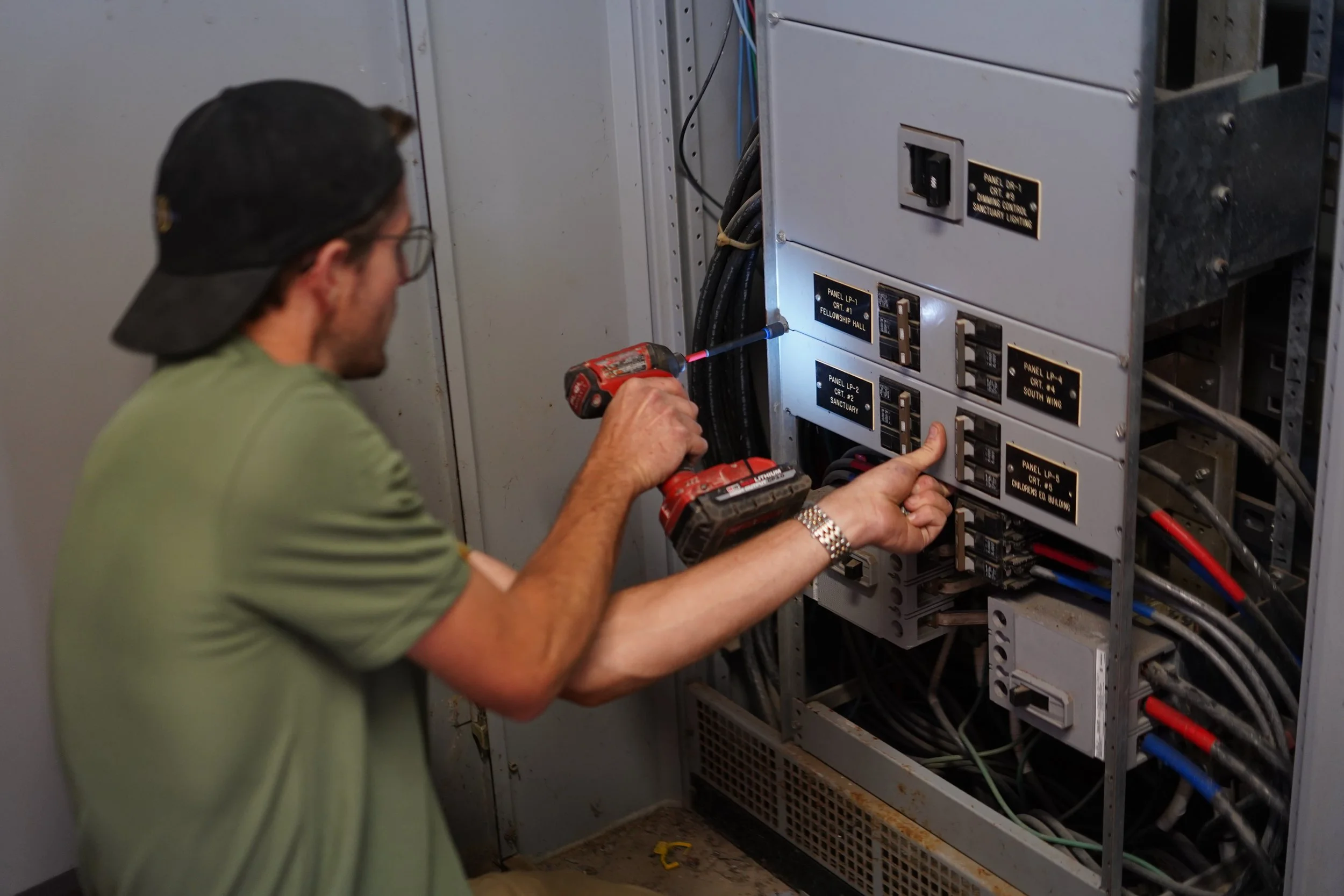 A man wearing a black cap, glasses, and a green t-shirt is working on an electrical panel with a screwdriver. He is using a cordless drill for assistance. The panel contains various switches and wires, with black labels and gold lettering.
