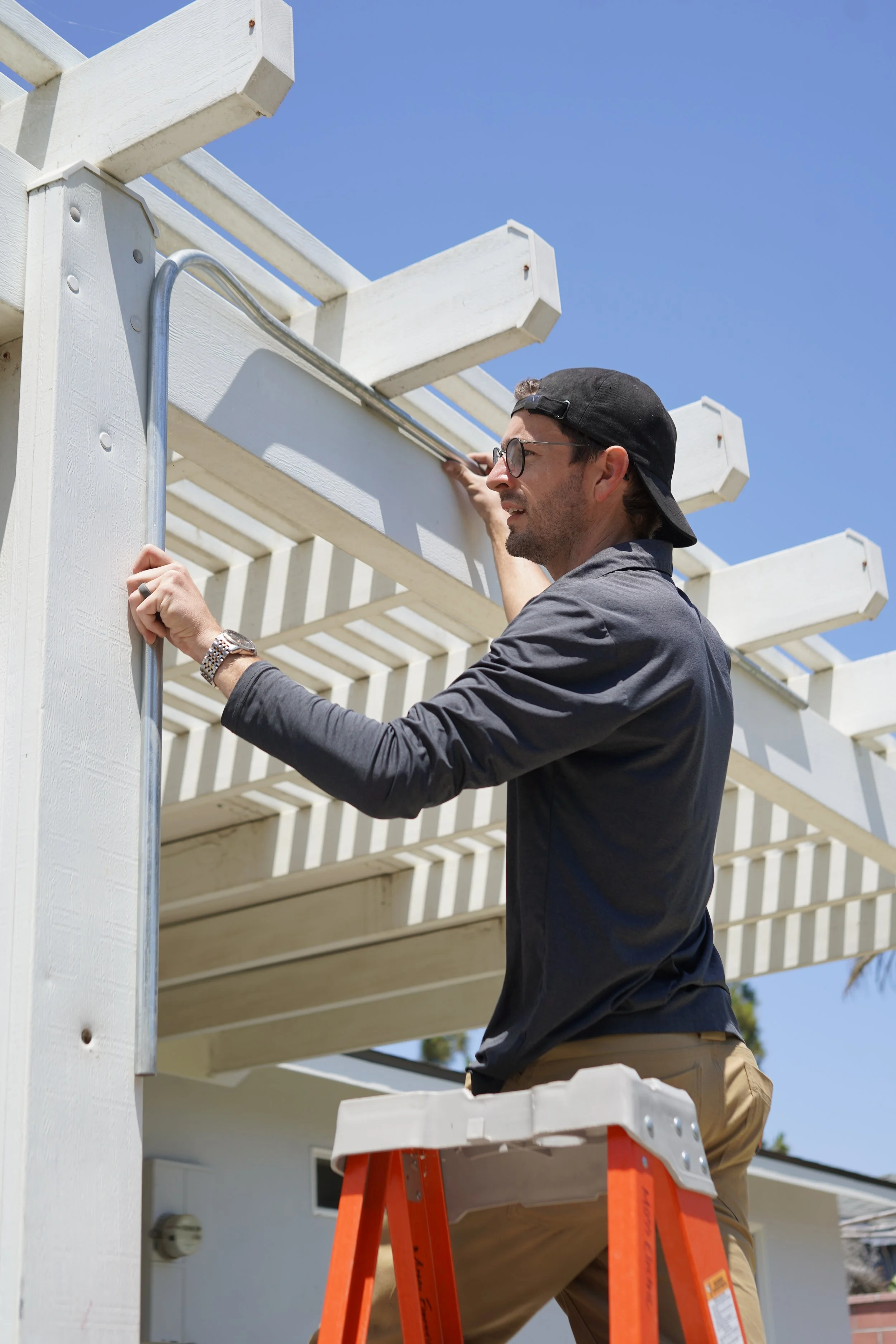 A man wearing glasses, a black cap, a long sleeve shirt, and khaki pants standing on an orange ladder, working on a white wooden structure outdoors under a clear blue sky.