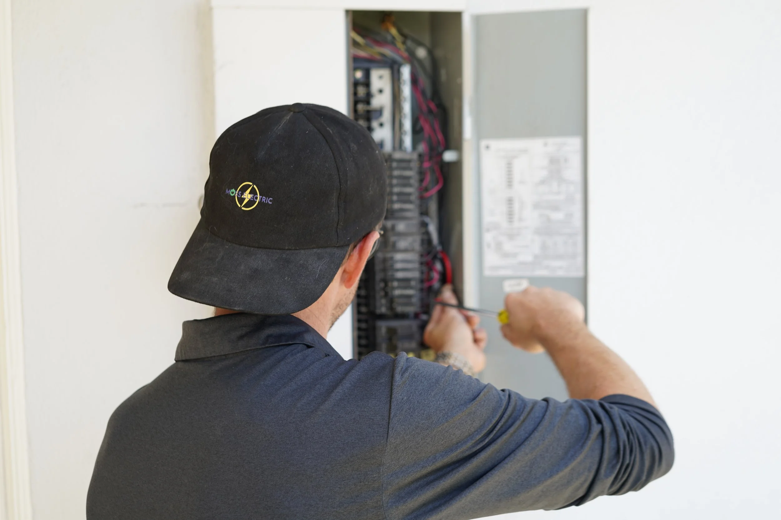 Technician working on an electrical panel, wearing a black cap with a company logo, using a screwdriver.