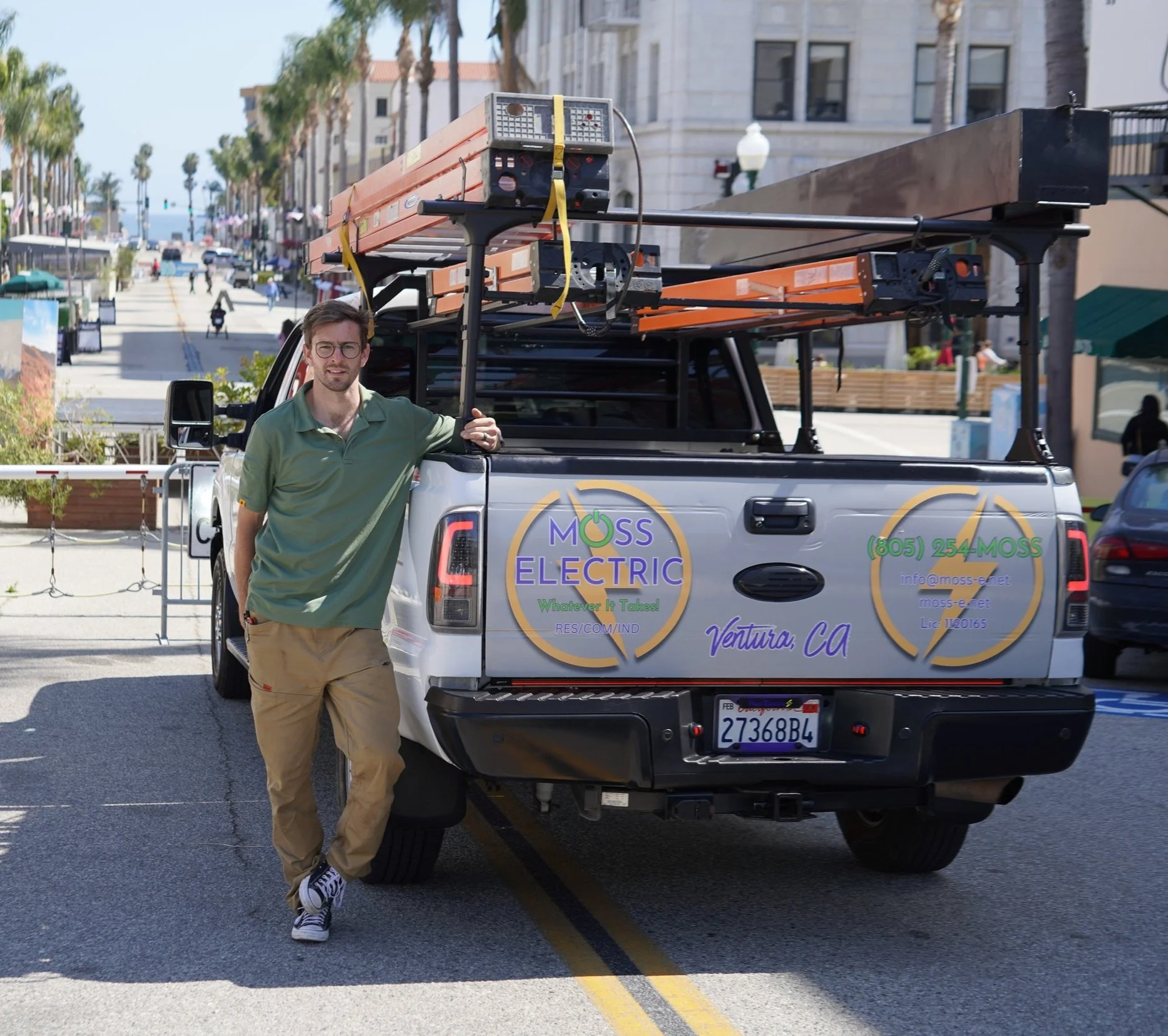 Man standing next to a Moss Electric service truck on a sunny street in Ventura, California, with a work ladder on the truck bed.