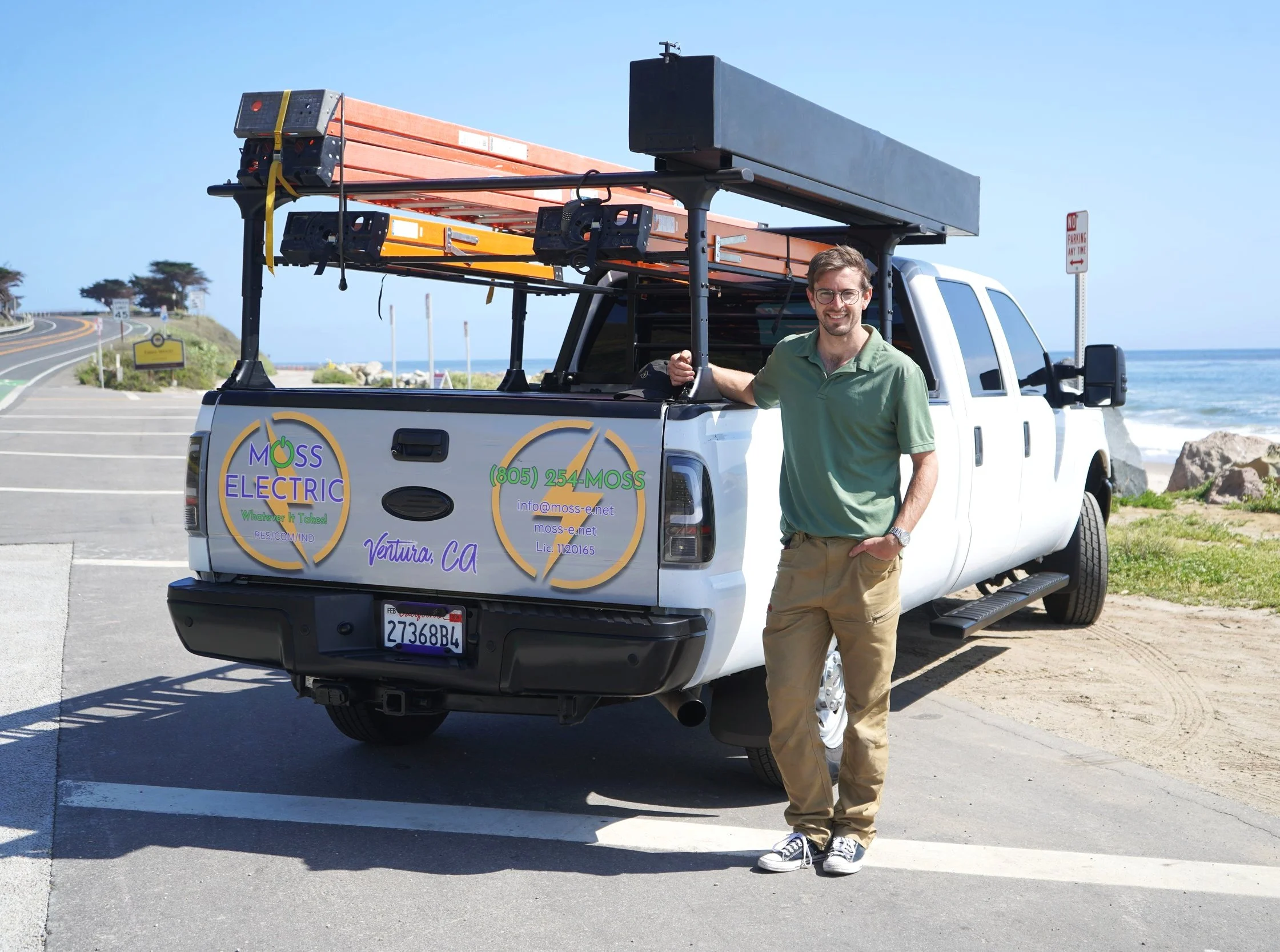 A man standing in front of a white pickup truck parked near the beach. The truck has a sign for 'MOSS ELECTRIC' with contact information, and the background shows the ocean, rocks, and a clear blue sky.