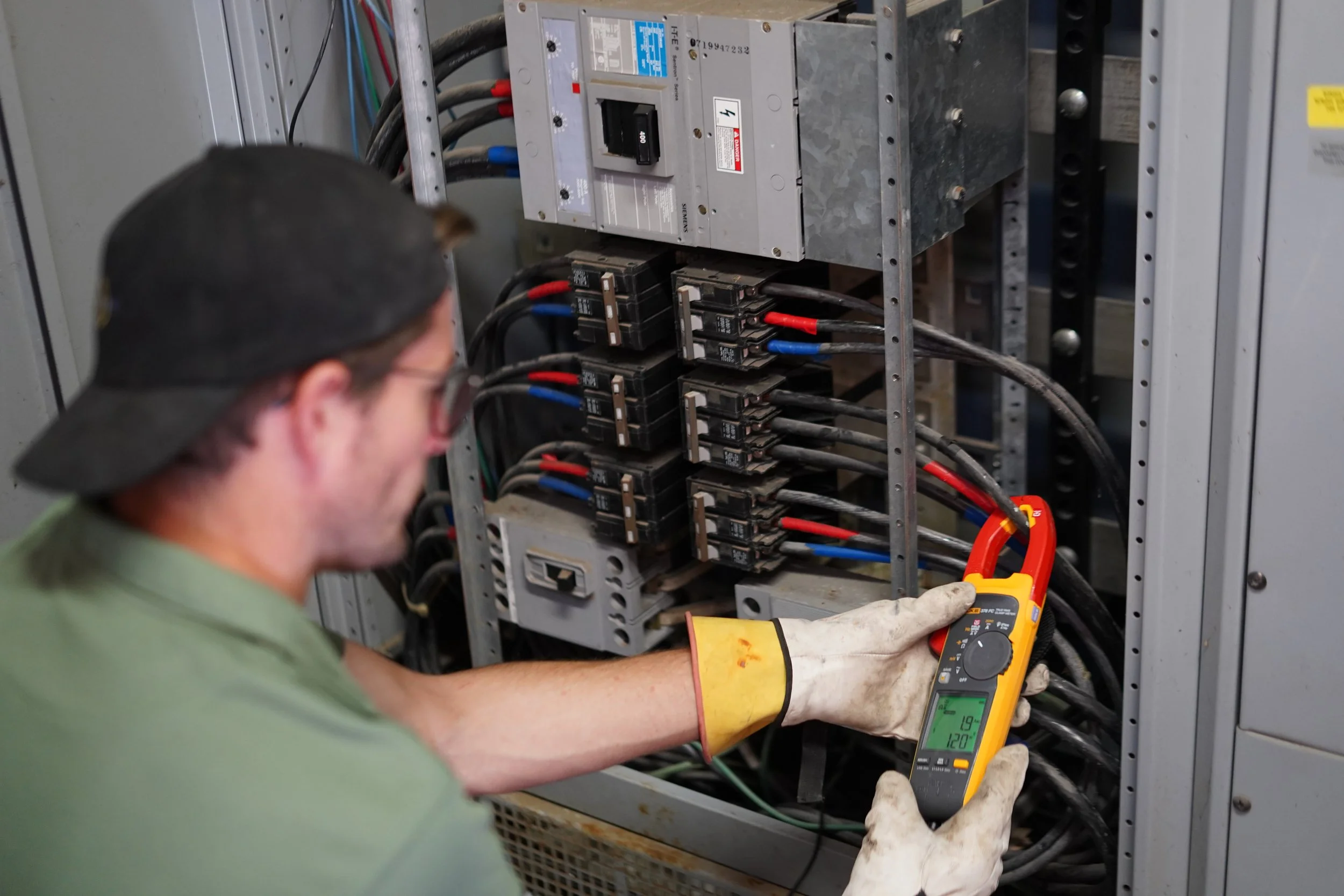 A technician wearing gloves and glasses is using a multimeter to test electrical components inside an open electrical panel.