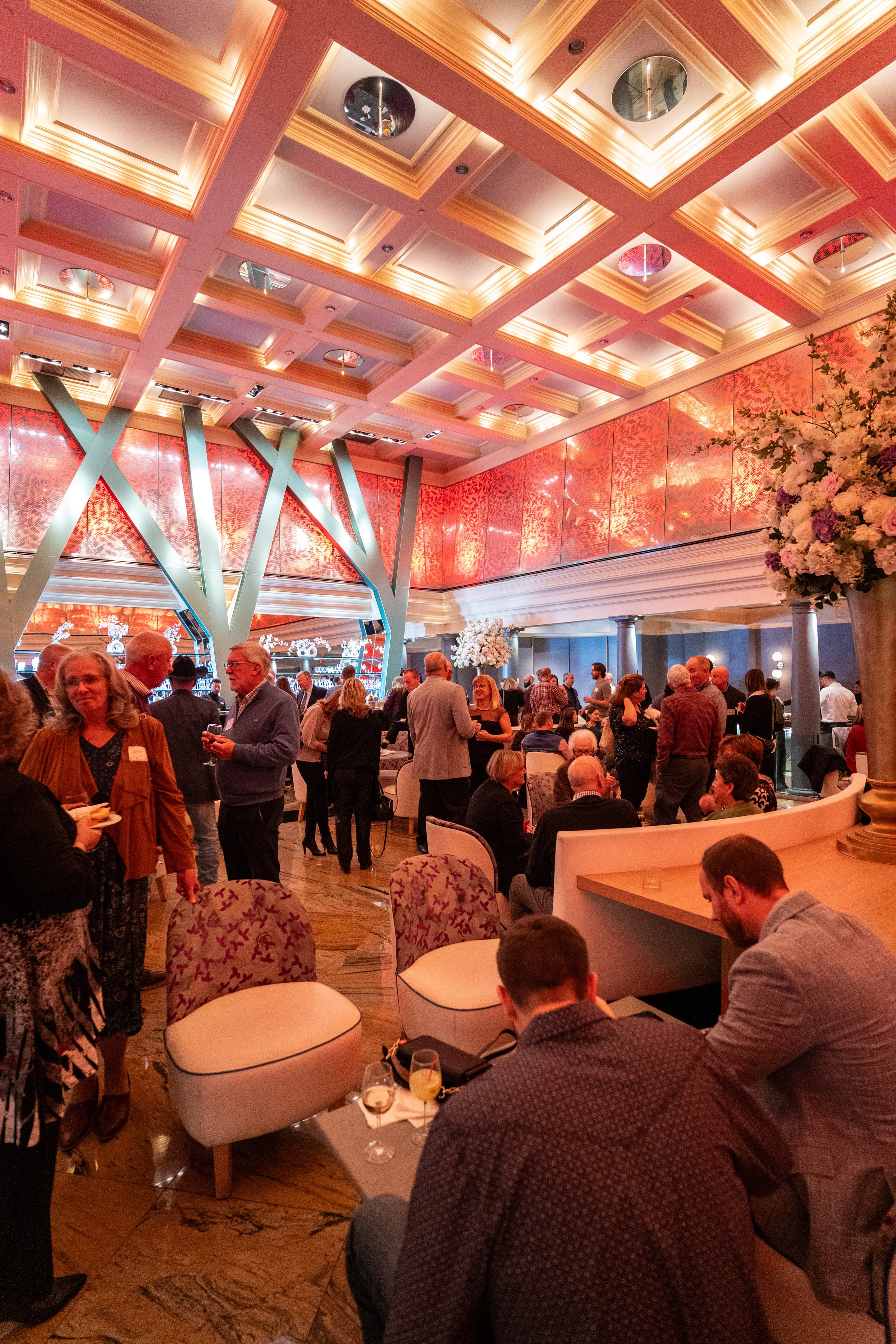 Interior of a bustling event space with a high, decorated ceiling, large floral arrangements, and many people socializing and seated at tables.