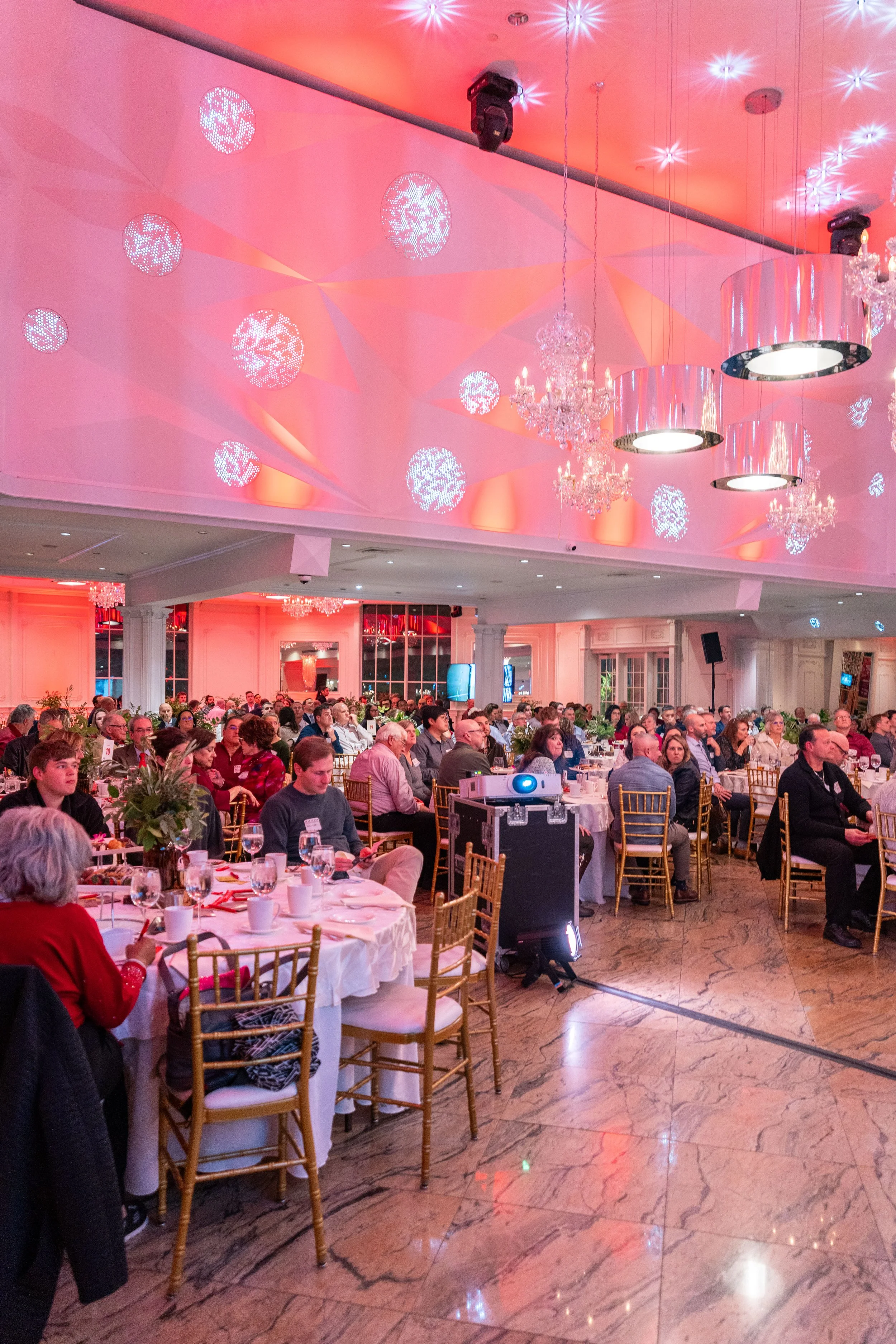 A large banquet hall filled with people sitting at decorated round tables, illuminated with pink and white lighting, chandeliers, and ceiling spotlights.
