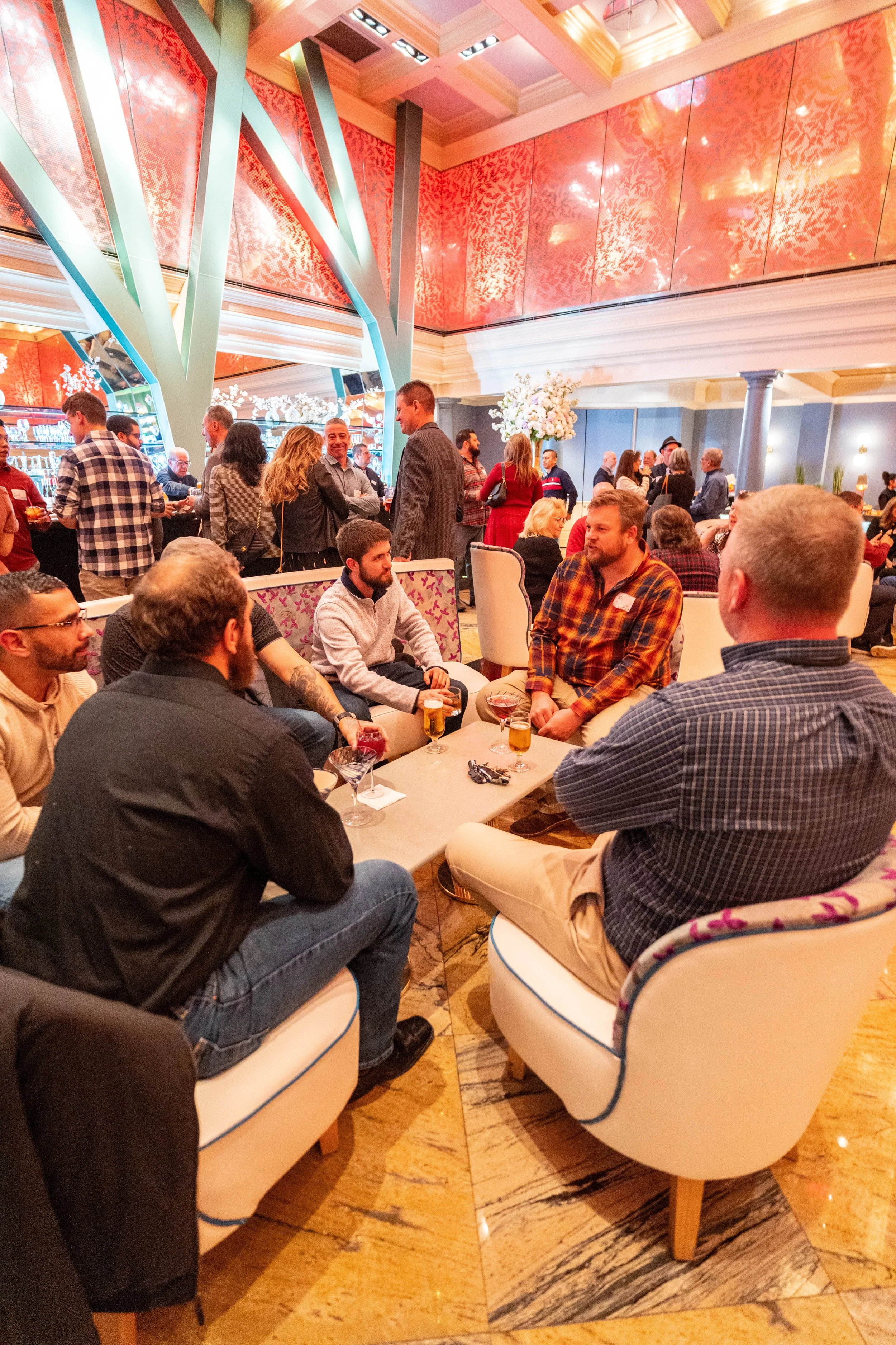 Indoor social gathering with people conversing and enjoying drinks in a brightly decorated venue with pink and green accents, floral arrangements, and a lively atmosphere.