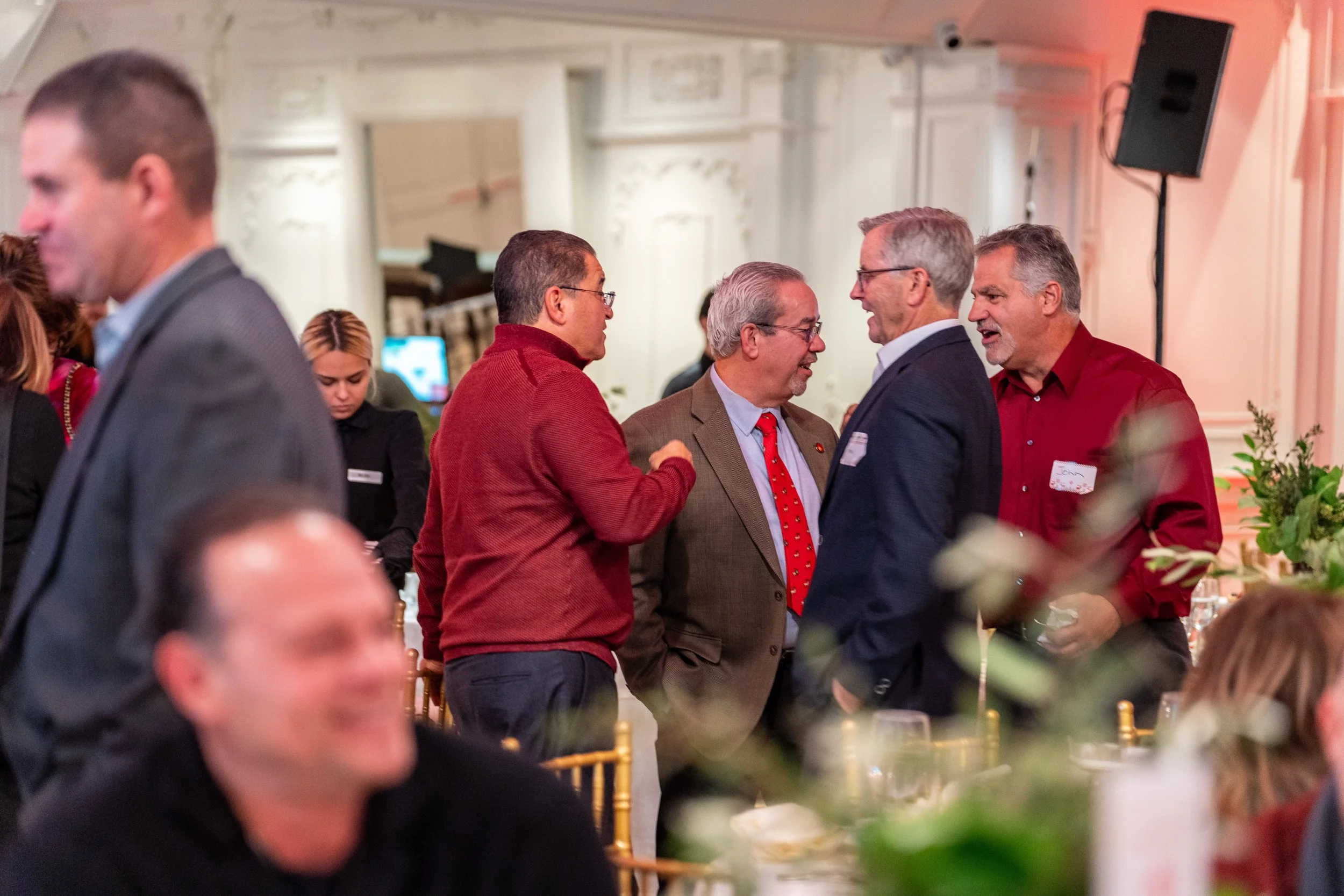 Group of five men engaged in conversation at a formal event, some wearing suits and others in business casual attire, in a well-lit banquet room with decorated walls and tables with floral arrangements.
