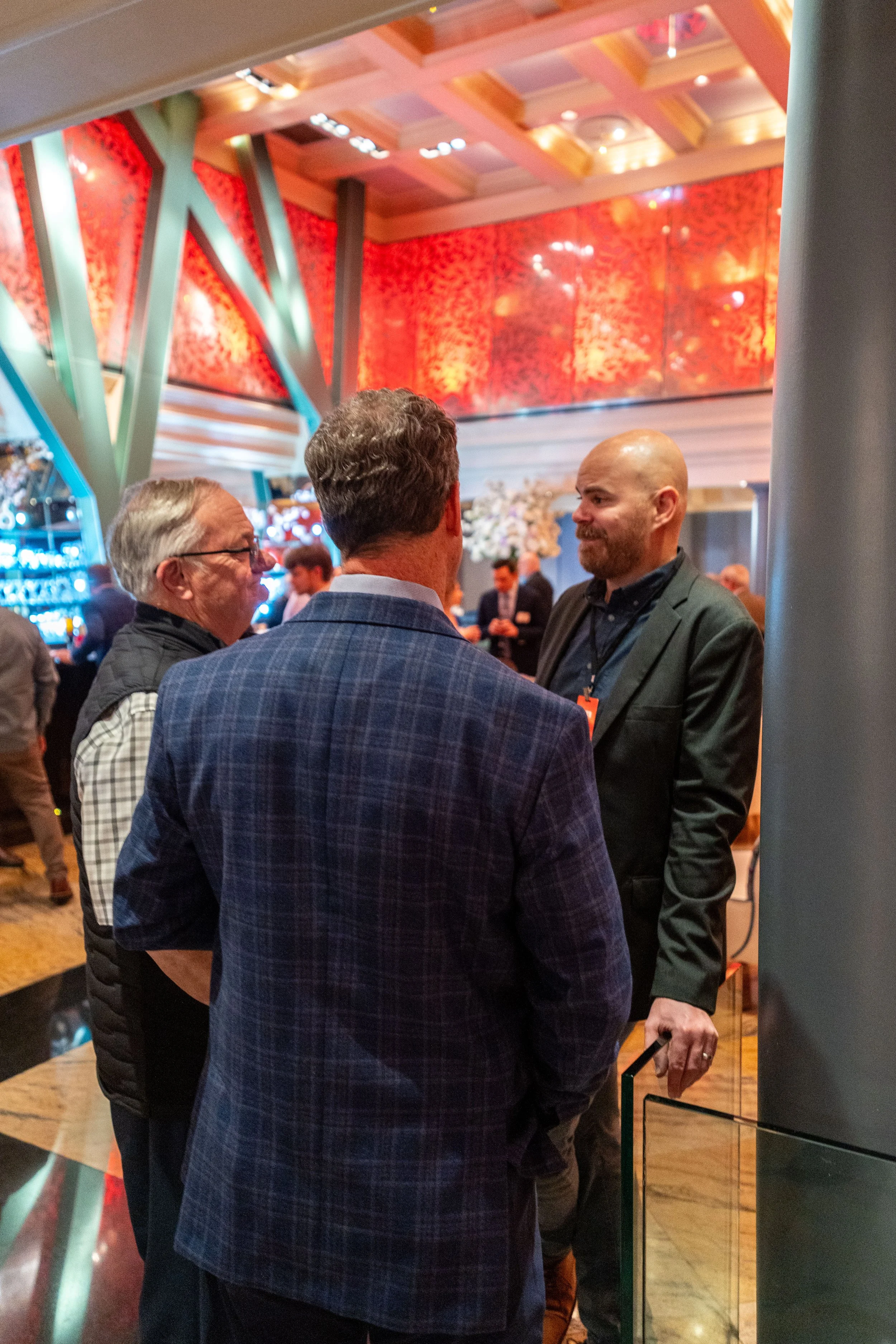 Three men engaged in conversation at a formal event with a modern interior and warm lighting.