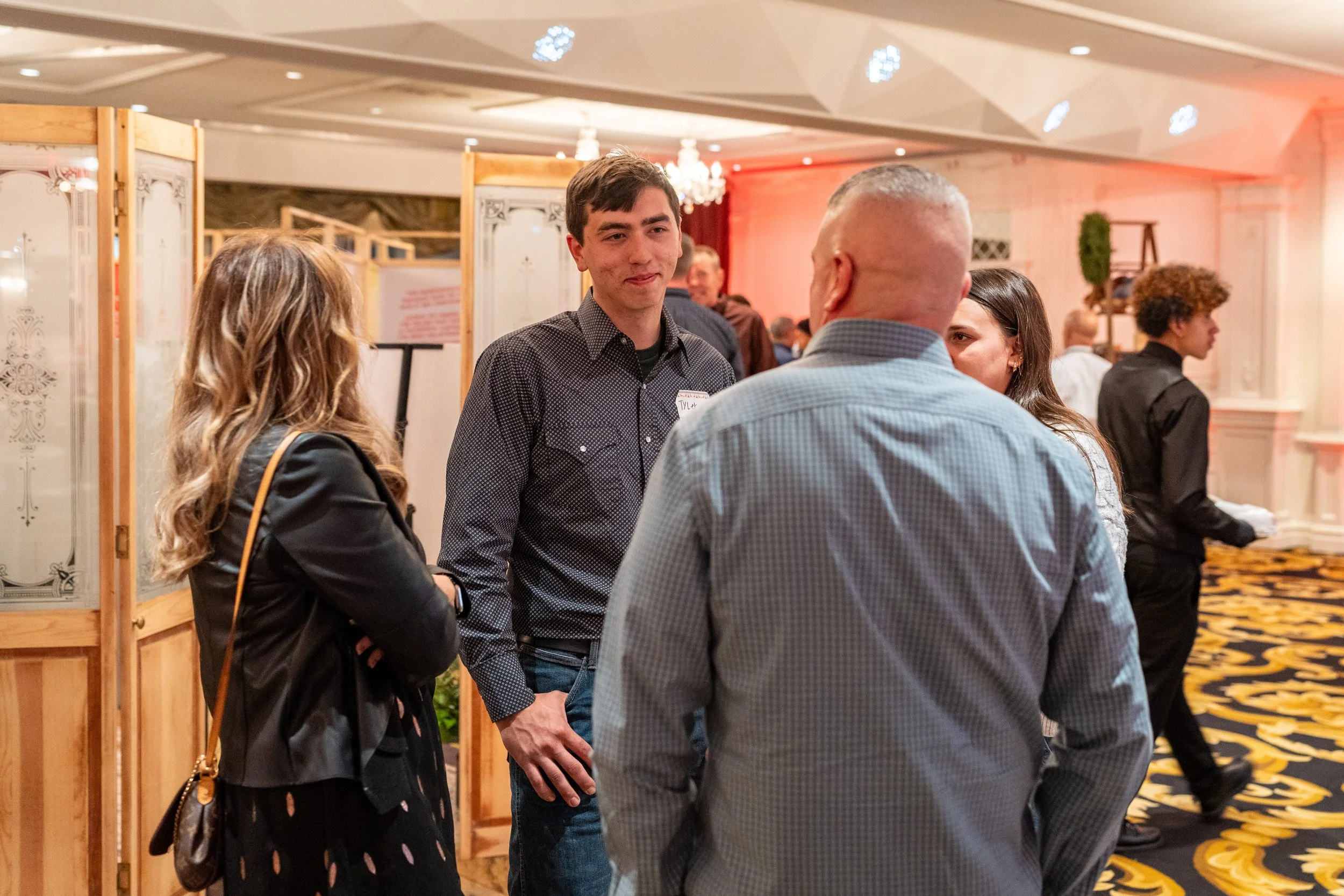Group of four people talking in a conference or networking event, with a woman on the left, a man in the center, and two people on the right, one further back.