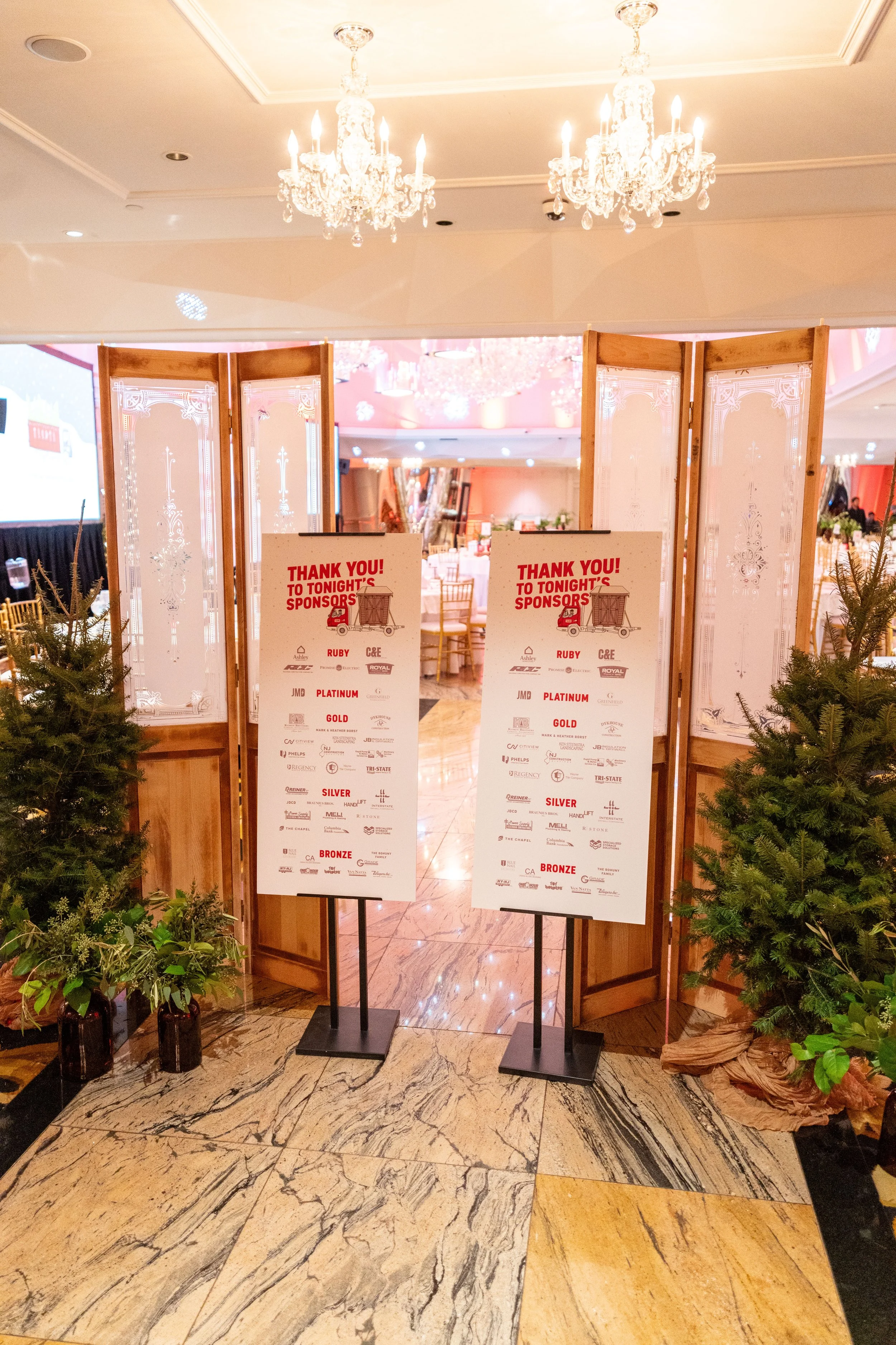 Event entrance with two sponsor thank you signs, potted plants, and a background of a decorated banquet hall with chandeliers.
