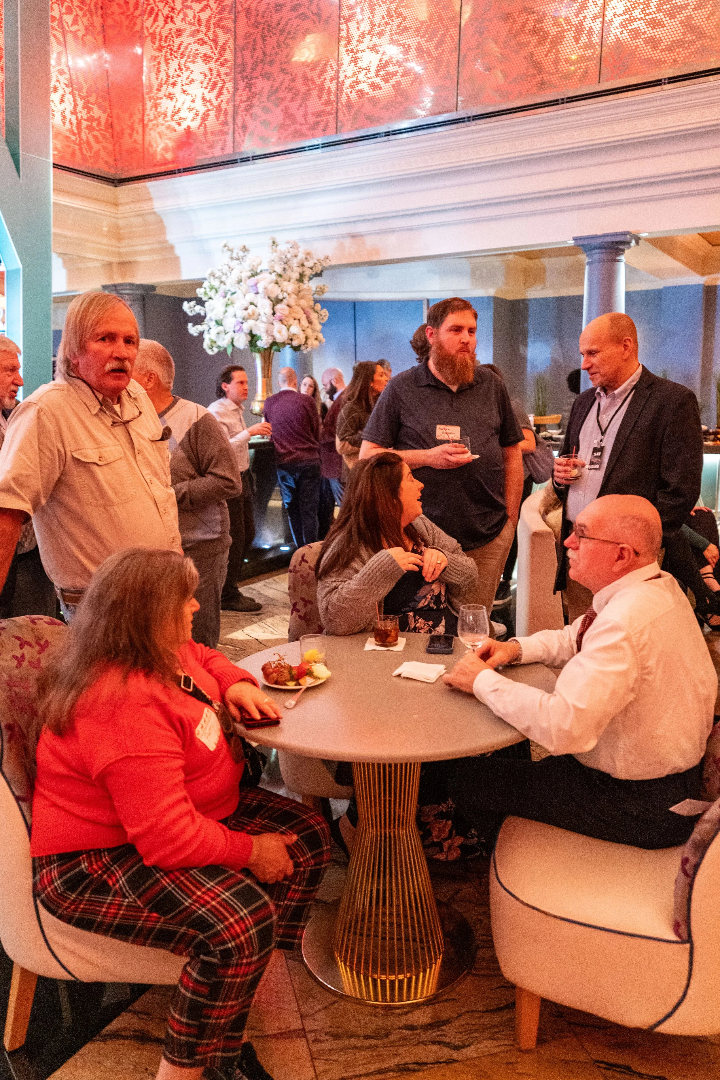 People socializing at a banquet or conference with drinks and food in a decorated venue with floral arrangements.