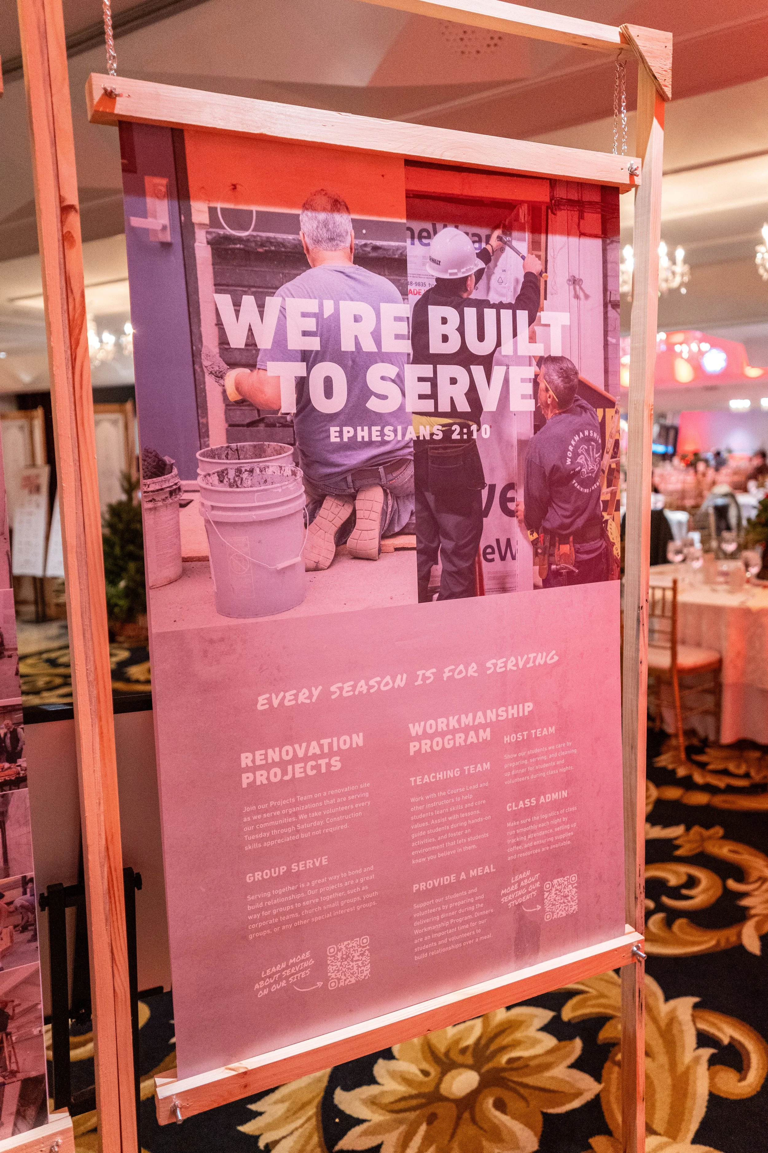 A sign at a construction-themed event displays the message 'We're built to serve' with a photo of workers and text about renovation projects, work program, teaching team, class admin, and providing meals.