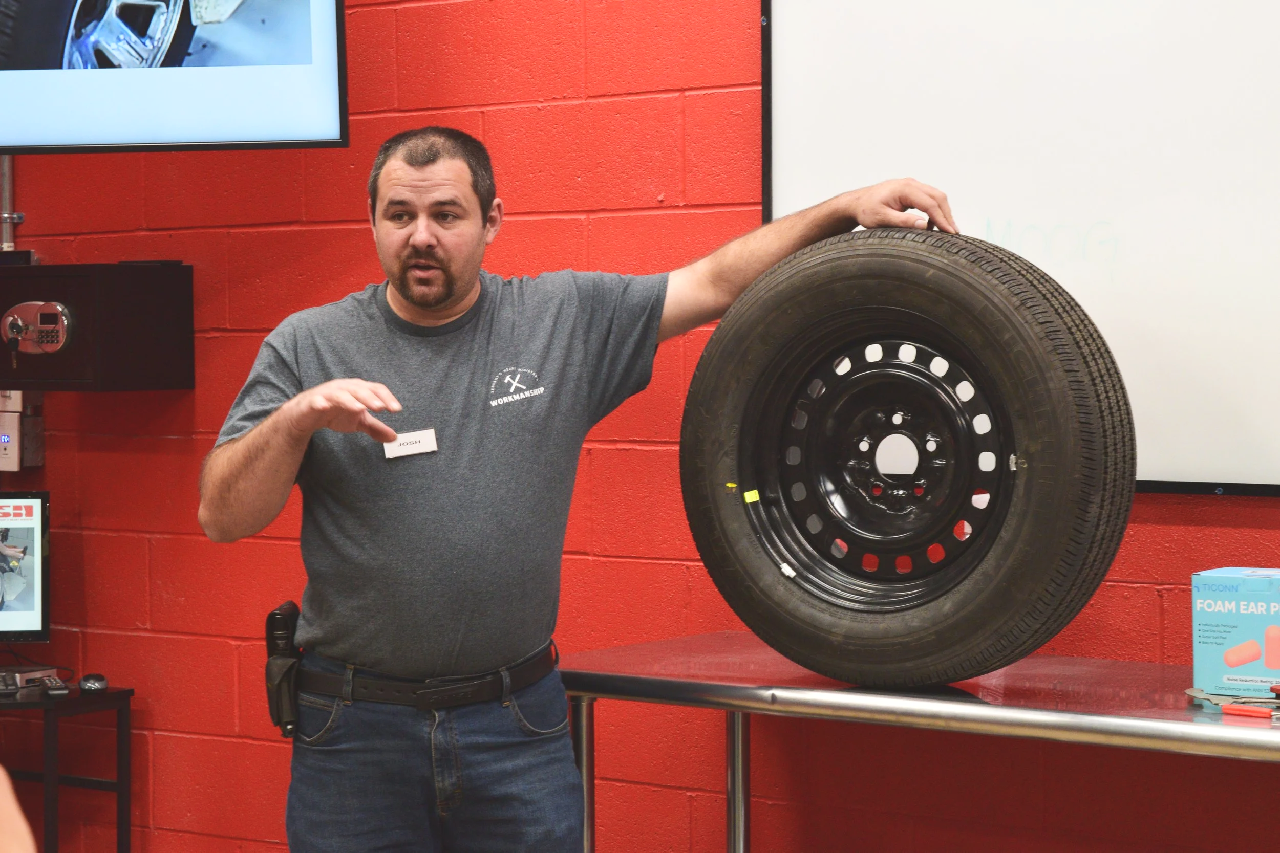 A man explaining something with his hand, standing next to a car tire on a table, in a room with red walls and a whiteboard.