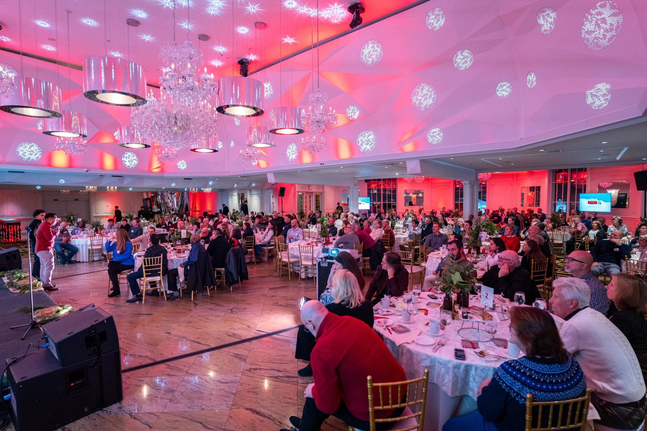 A large banquet hall decorated with pink and purple lighting, chandeliers, and snowflake patterns projected onto the ceiling during a formal event with numerous round tables, guests, and a small stage area.