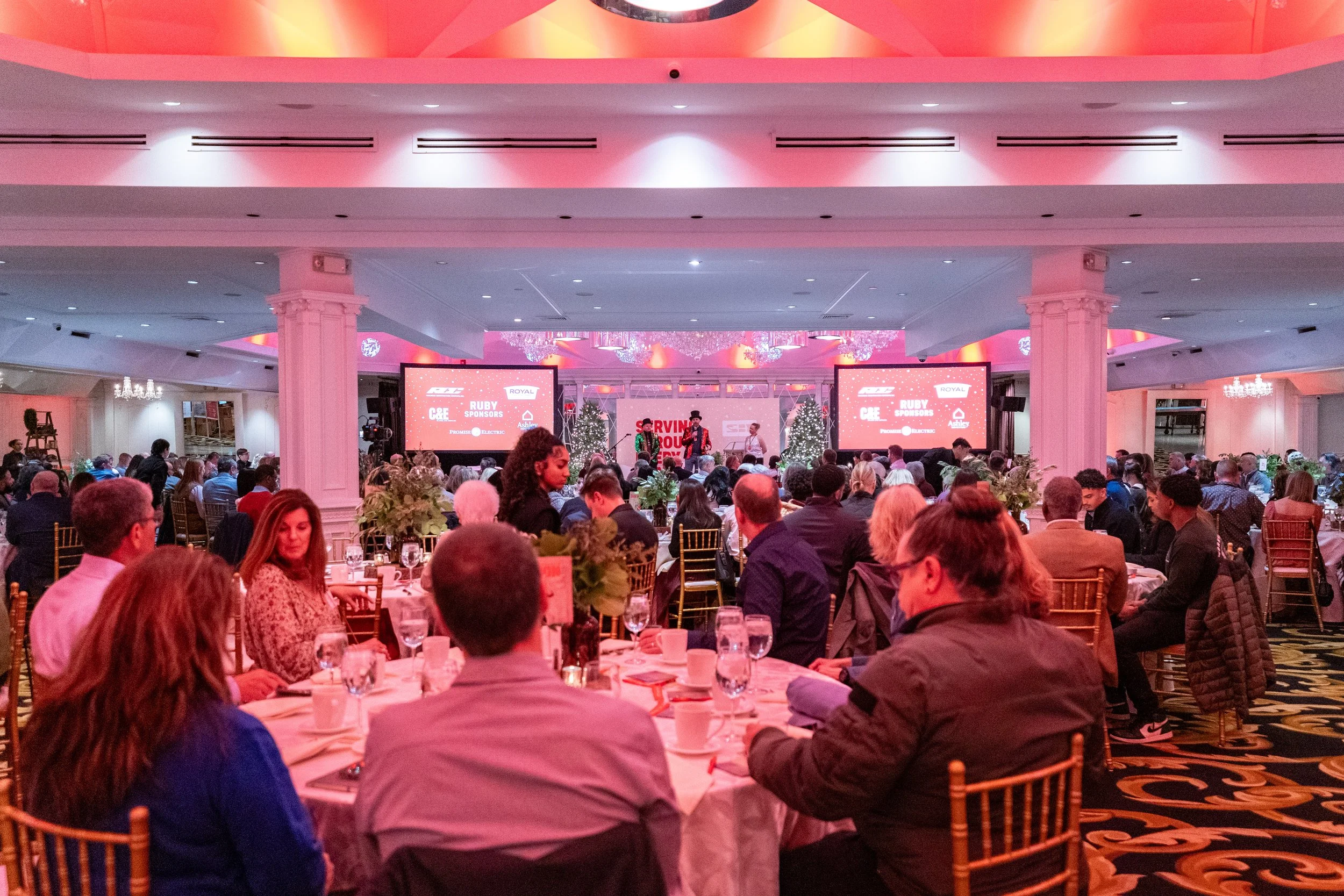 A large banquet hall filled with people seated at decorated tables, watching a stage with two large screens and Christmas trees, pink and white lighting, and festive holiday decorations.