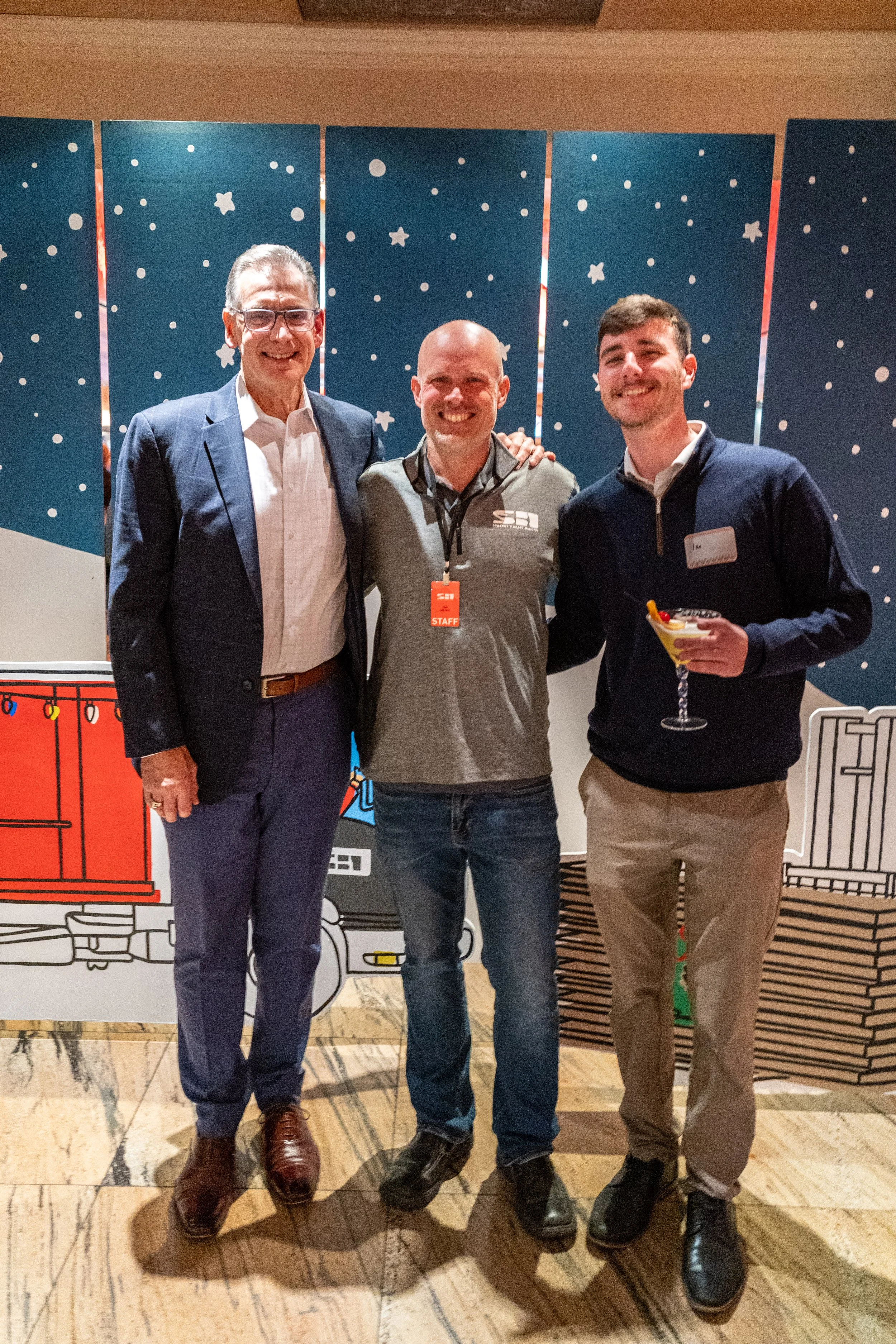 Three men standing together at a holiday party, smiling, with a festive holiday-themed backdrop behind them. The man on the left is wearing glasses, a dark blazer, white shirt, and brown shoes. The man in the middle is bald, wearing a gray polo with 