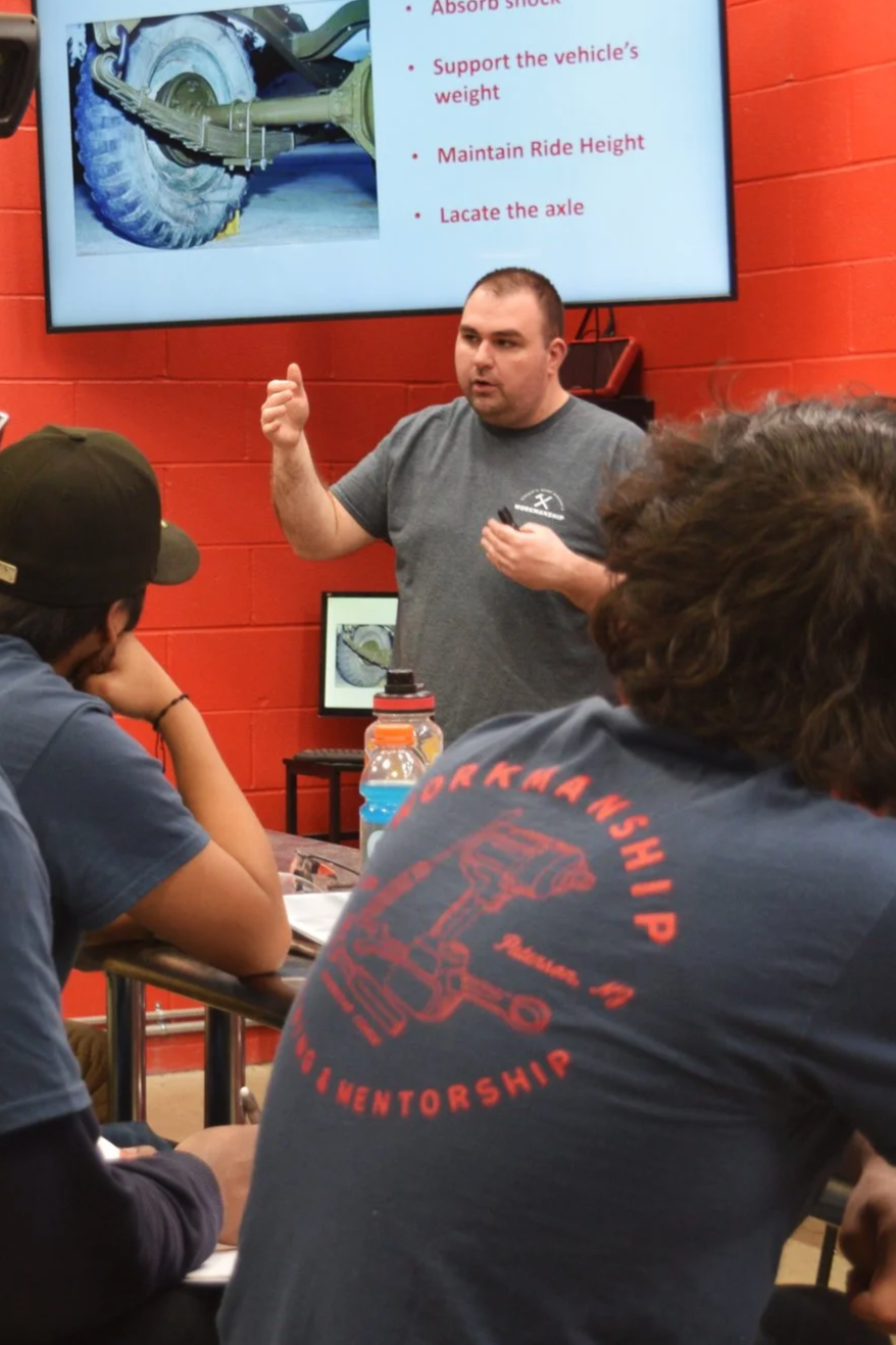 A man giving a presentation about vehicle axles in a classroom with students. A large screen behind him displays a diagram of a vehicle axle, with text listing steps like absorb shock and support the vehicle's weight. Students are seated at desks, listening and taking notes.