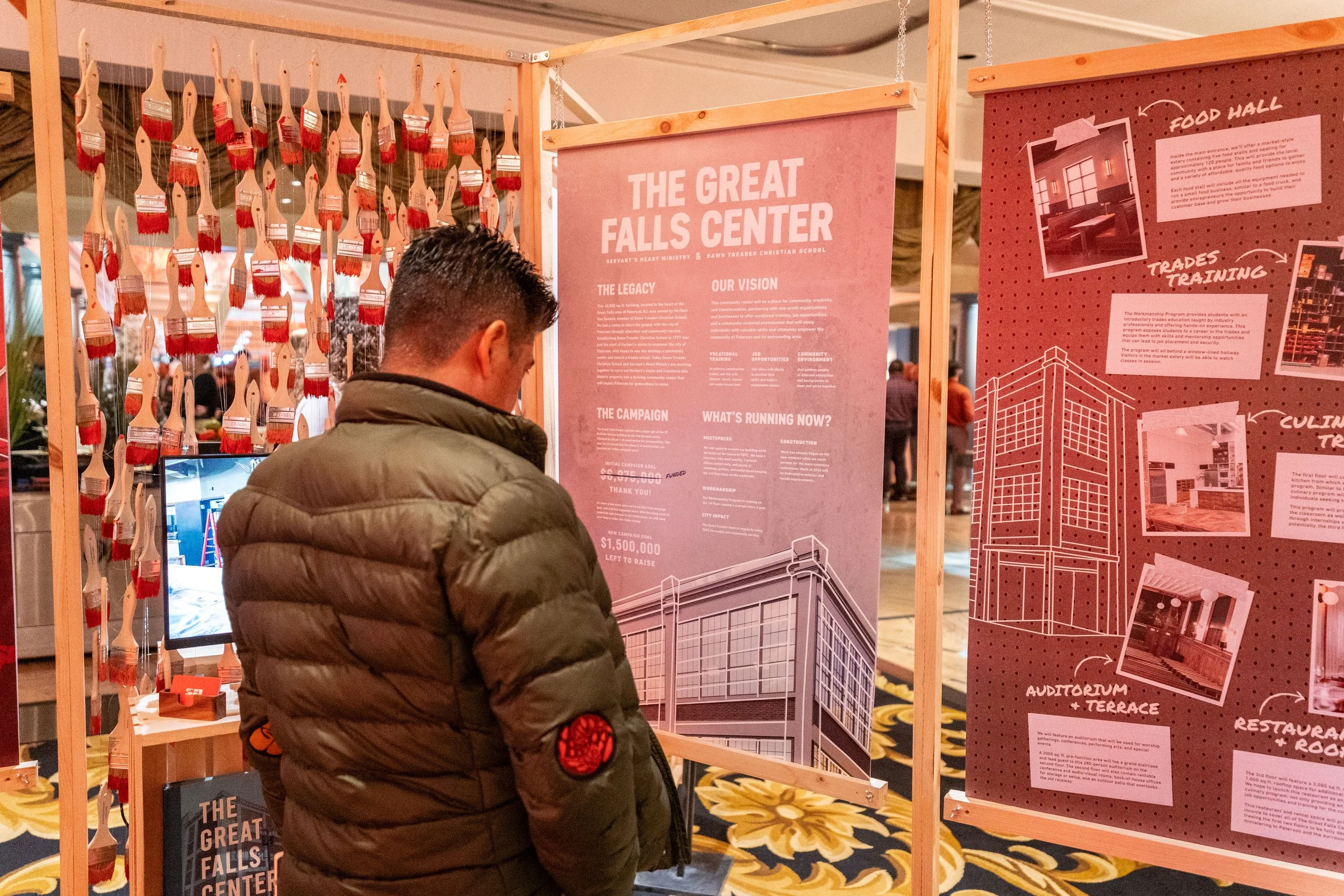 A person in a green puffer jacket looking at information boards about The Great Falls Center at an indoor event.