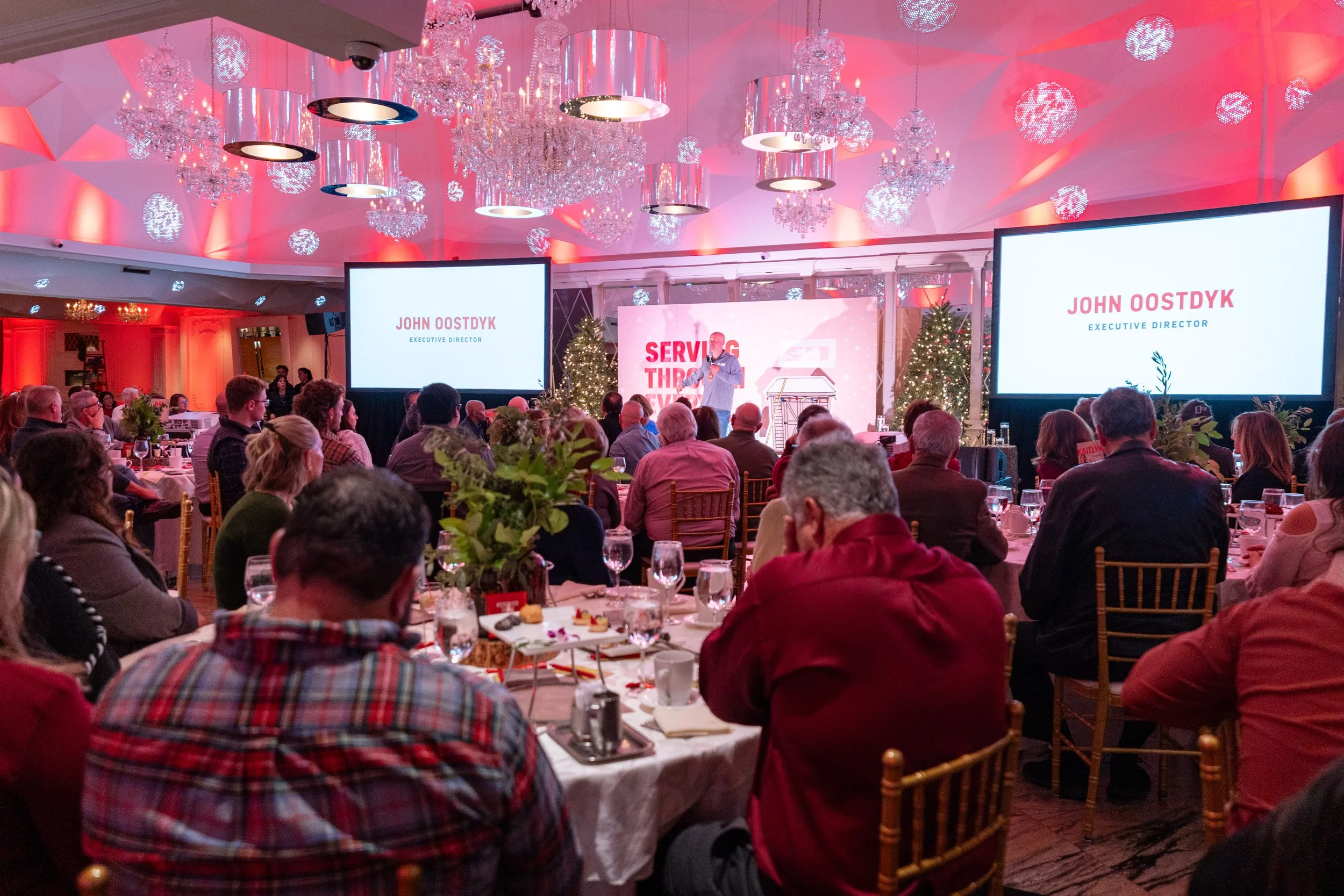 A conference room decorated with Christmas trees, chandeliers, and pink lighting. Audience seated at round tables watch a speaker on stage. Large screens display 'John Oostdyk, Executive Director' and a banner reads 'Serving Through'.