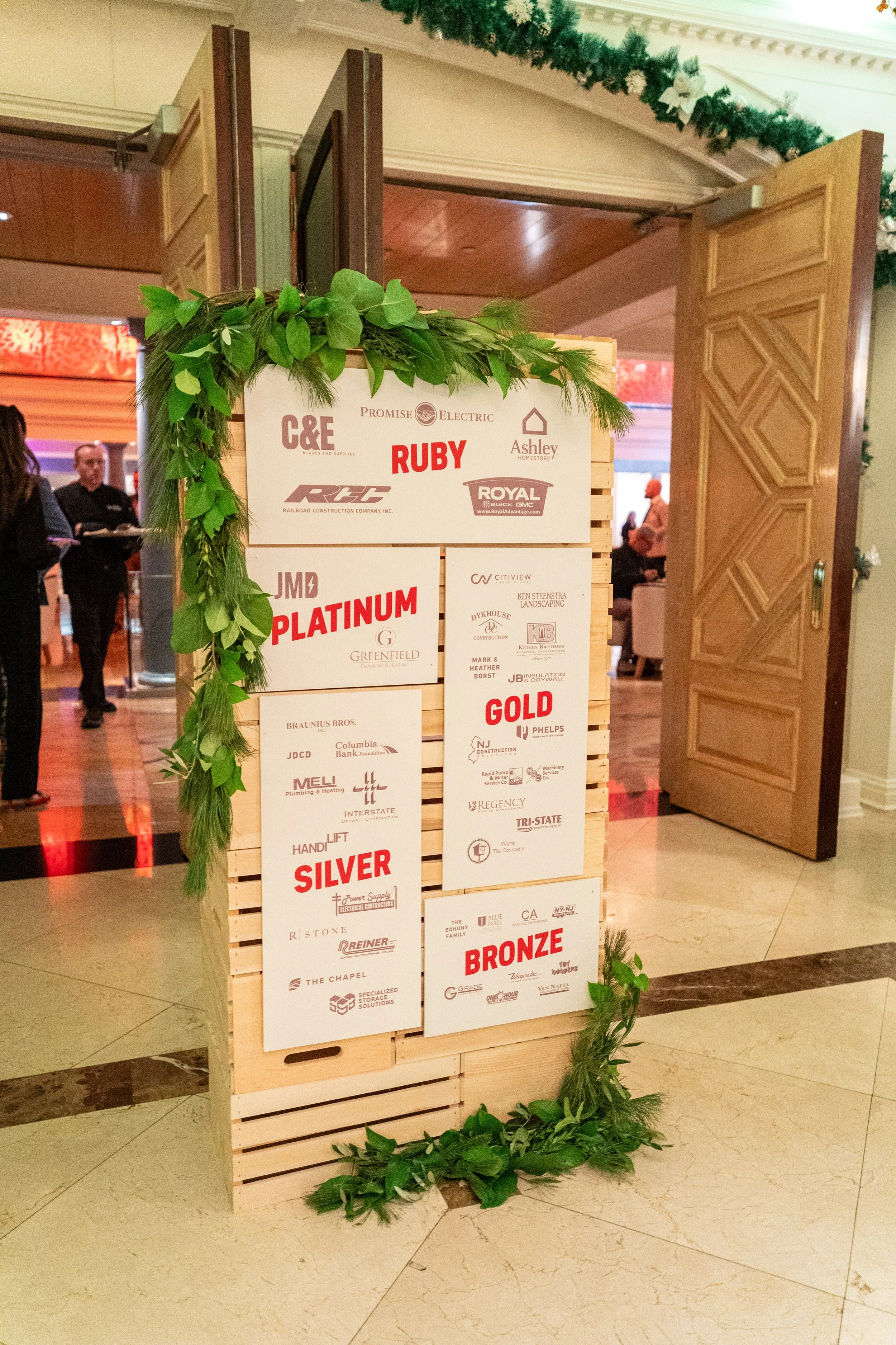 A wooden event sign decorated with green garland, displaying sponsors and awards for categories: Ruby, Platinum, Silver, Gold, and Bronze at a celebration or awards event.