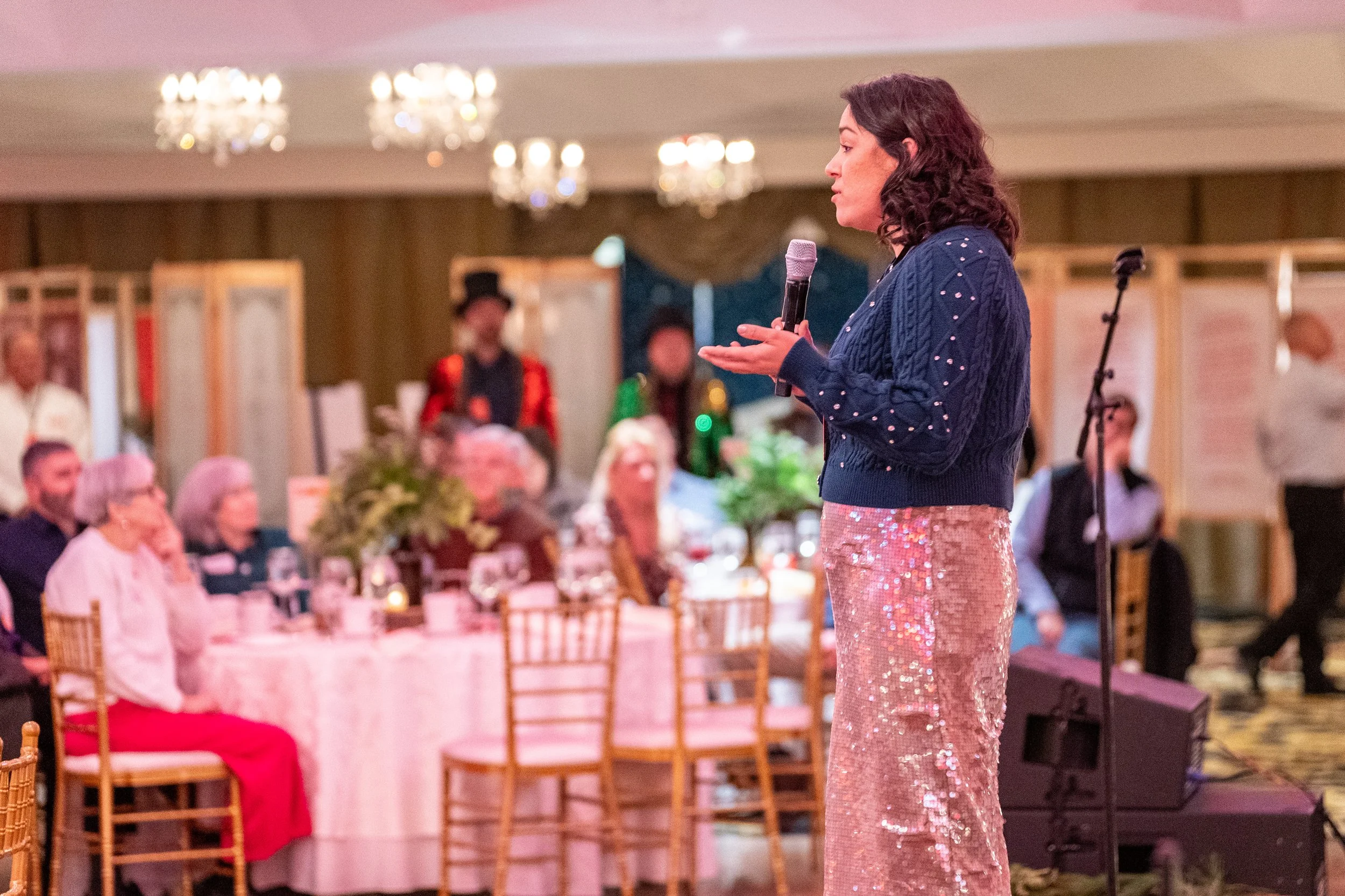 A woman with dark curly hair stands on a stage holding a microphone during an event. She is wearing a navy blue sweater with metallic embellishments and a long sequined skirt. In the background, there are seated guests at a round table with a floral 